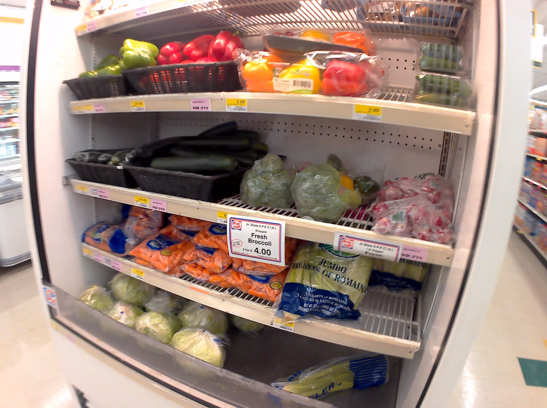 Refrigerated produce section at Discount Drug Mart with peppers, cucumbers, broccoli, and bags of vegetables.