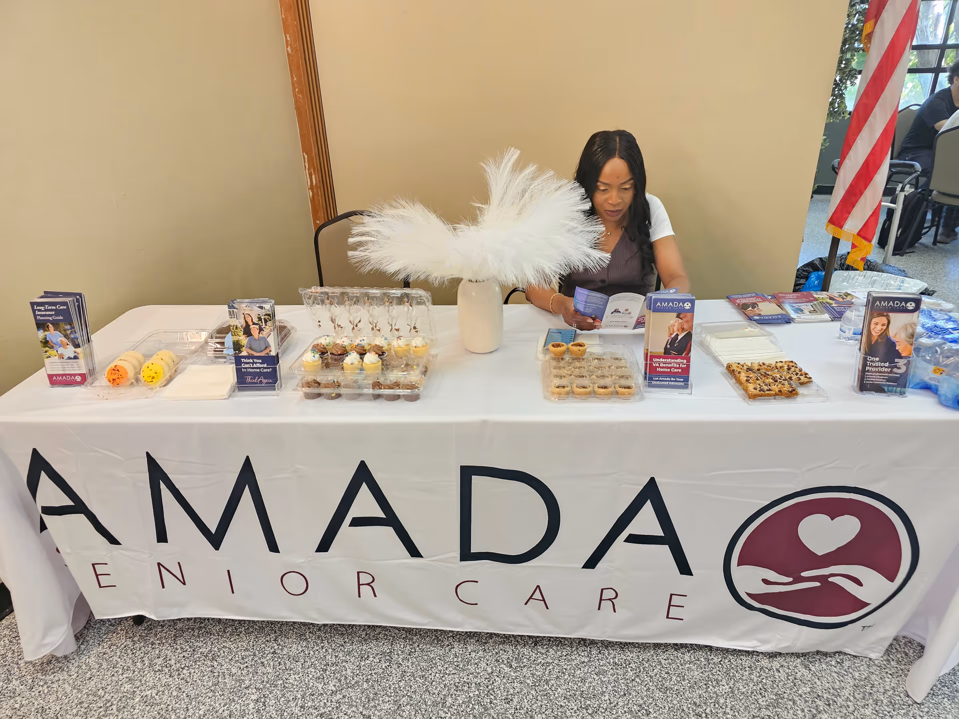 A staff member sits behind a promotional Amada Senior Care table with pastries in a lobby.