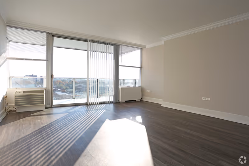 Bright empty living room with large windows and balcony doors, wood flooring.