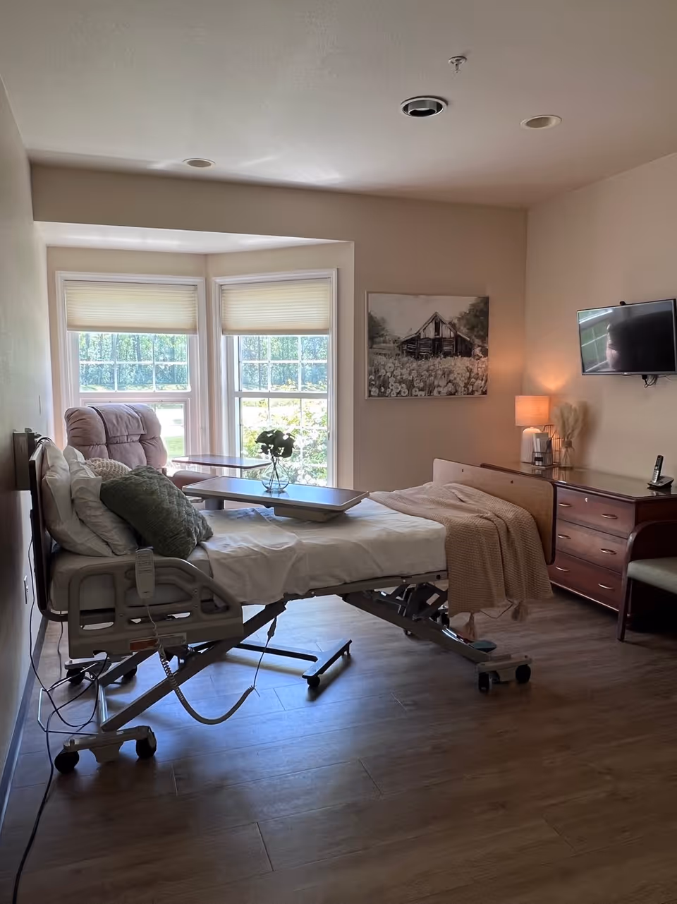 Hospital-style patient bed in a bright bedroom with windows and dresser.