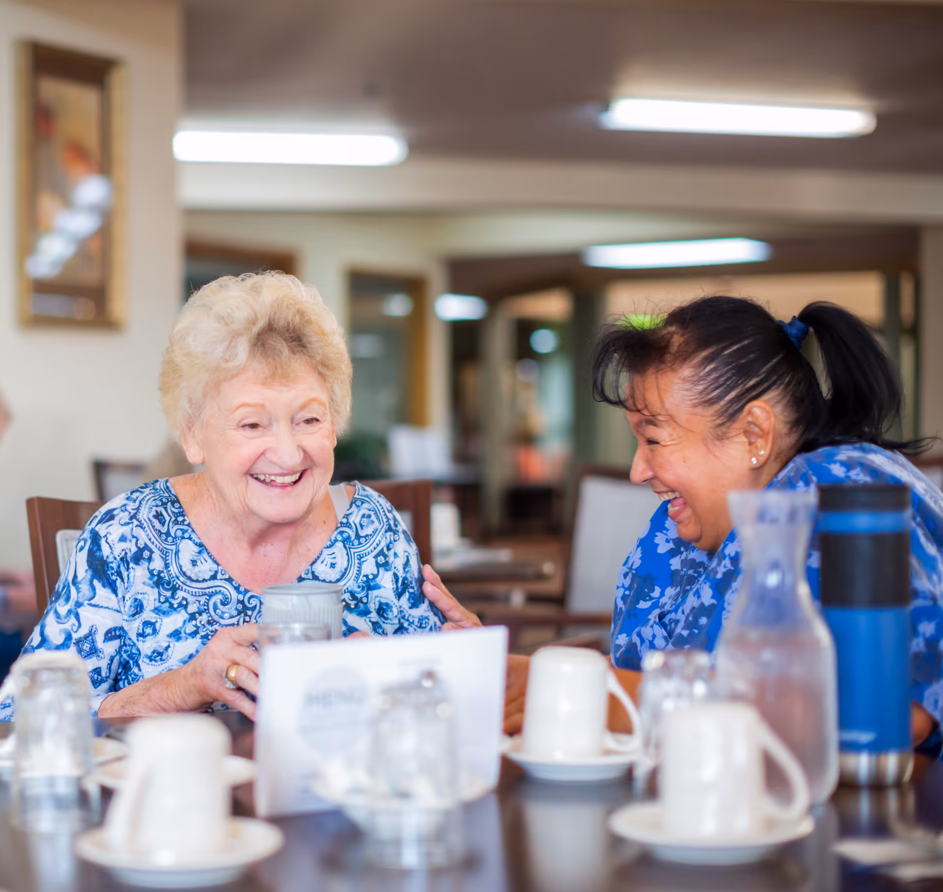 Two smiling seniors share a laugh at a dining table in a care facility.