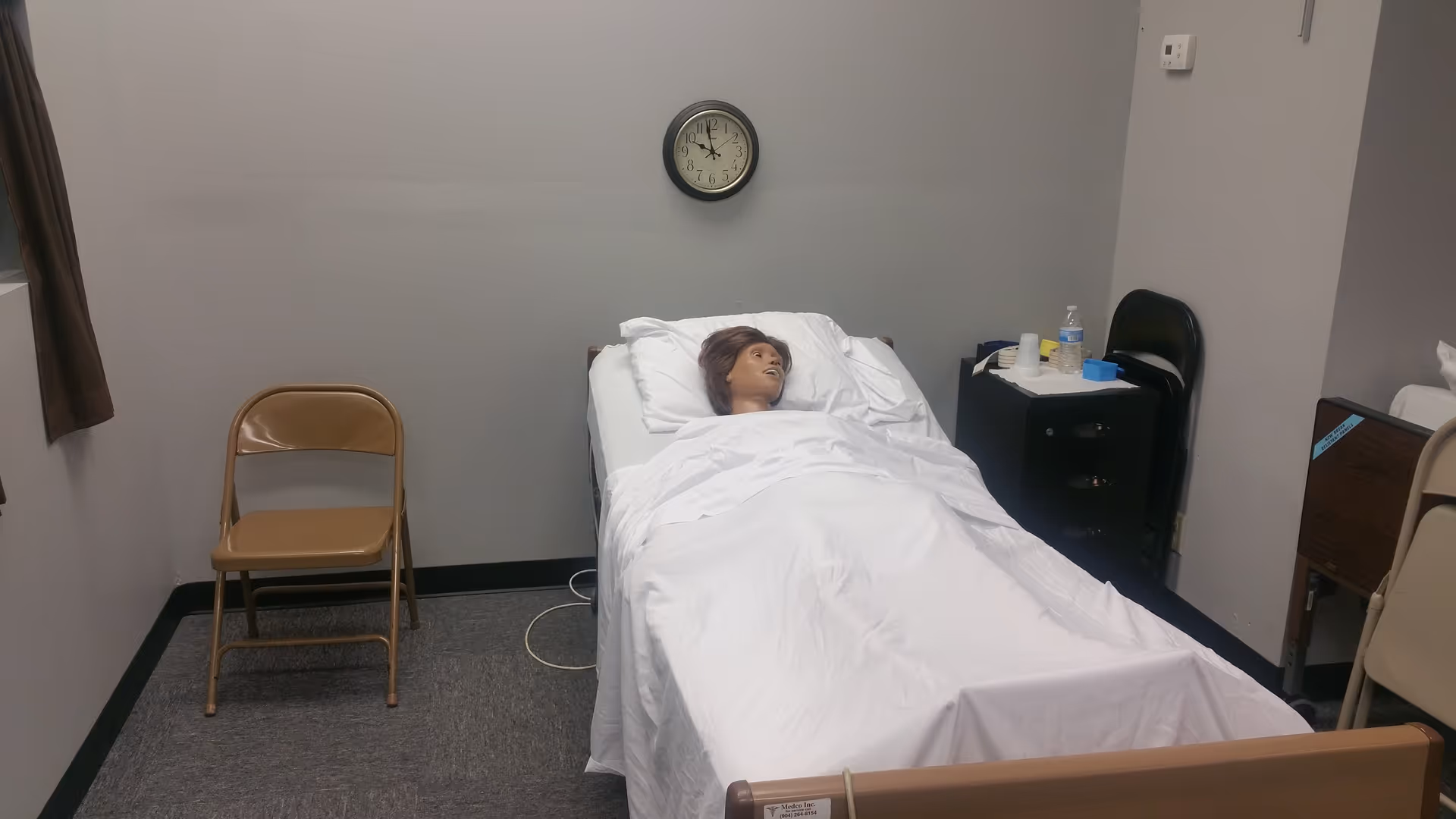Hospital-style bed with a mannequin patient covered by white sheets in a small training room.