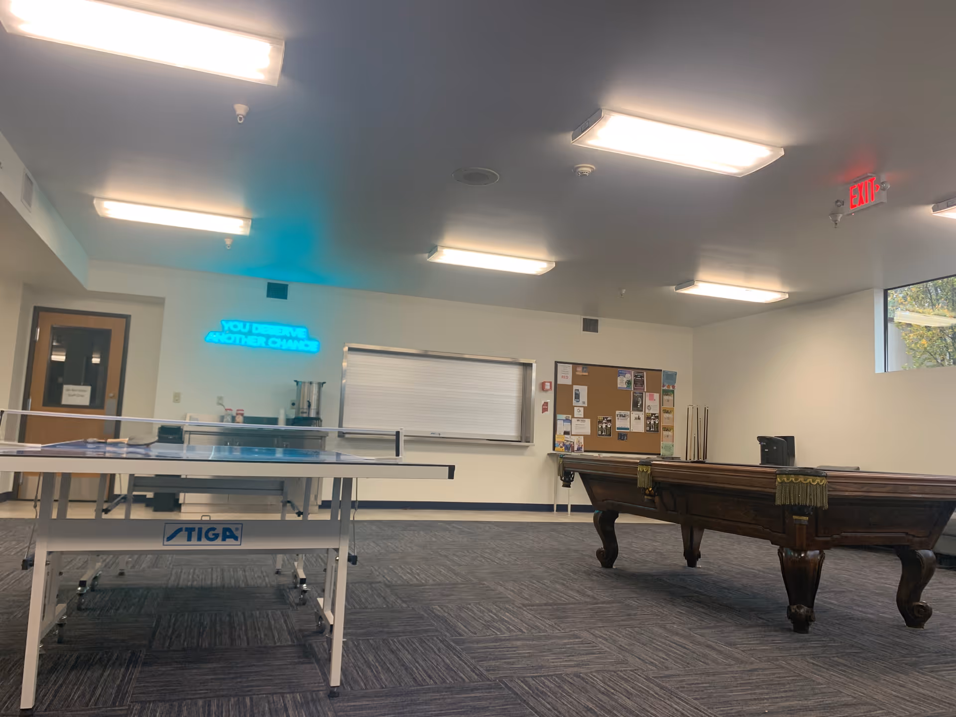 Indoor recreation room with ping-pong and billiards tables, corkboard, and bright overhead lighting.