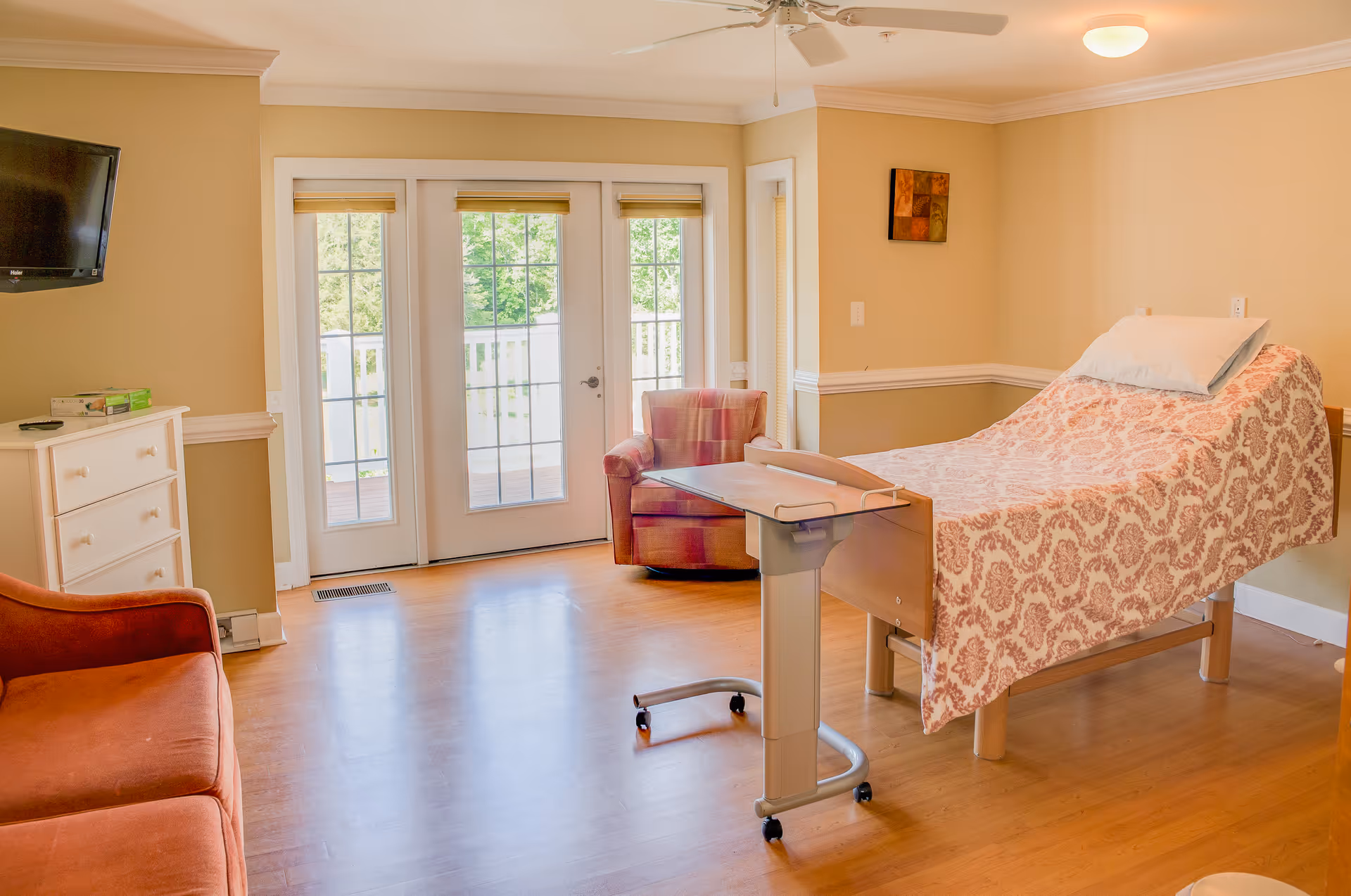 Hospice bedroom with a single bed, chair, dresser, TV, and French doors to outdoors.