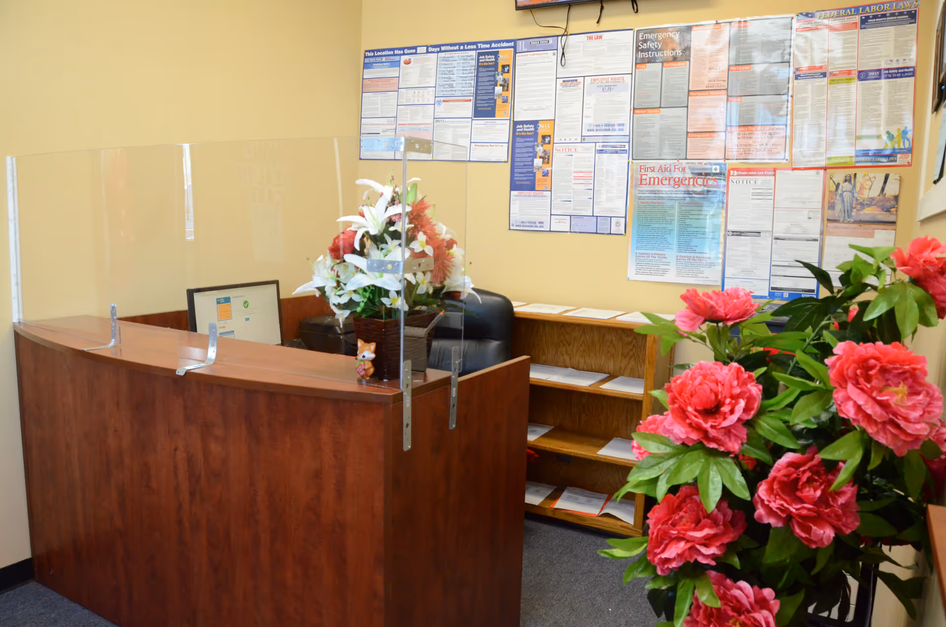 A wooden reception desk with a clear divider, notice boards, and pink flowers indoors.
