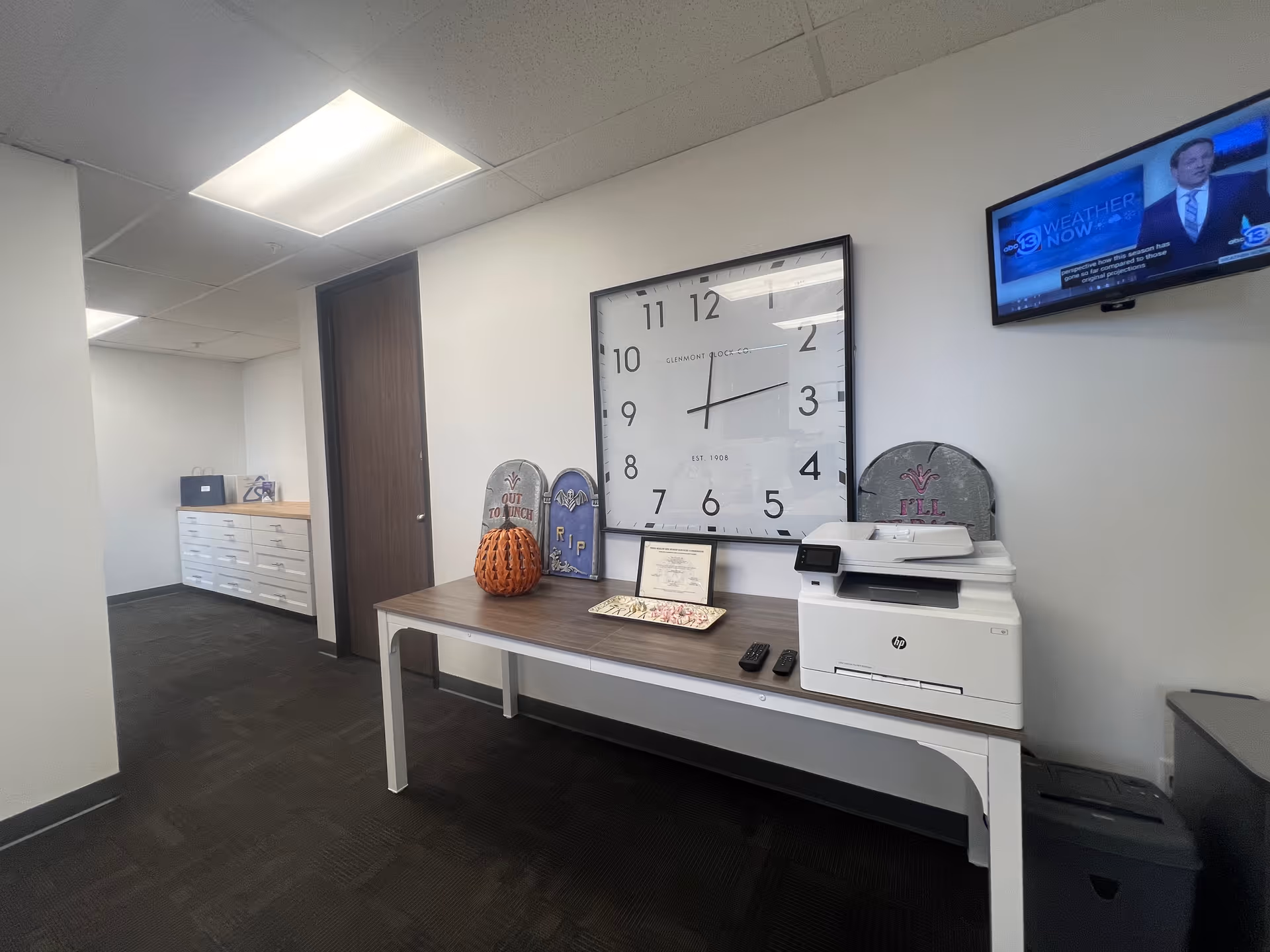Office lobby area with desk, large wall clock, printer, TV, and pumpkin decorations.