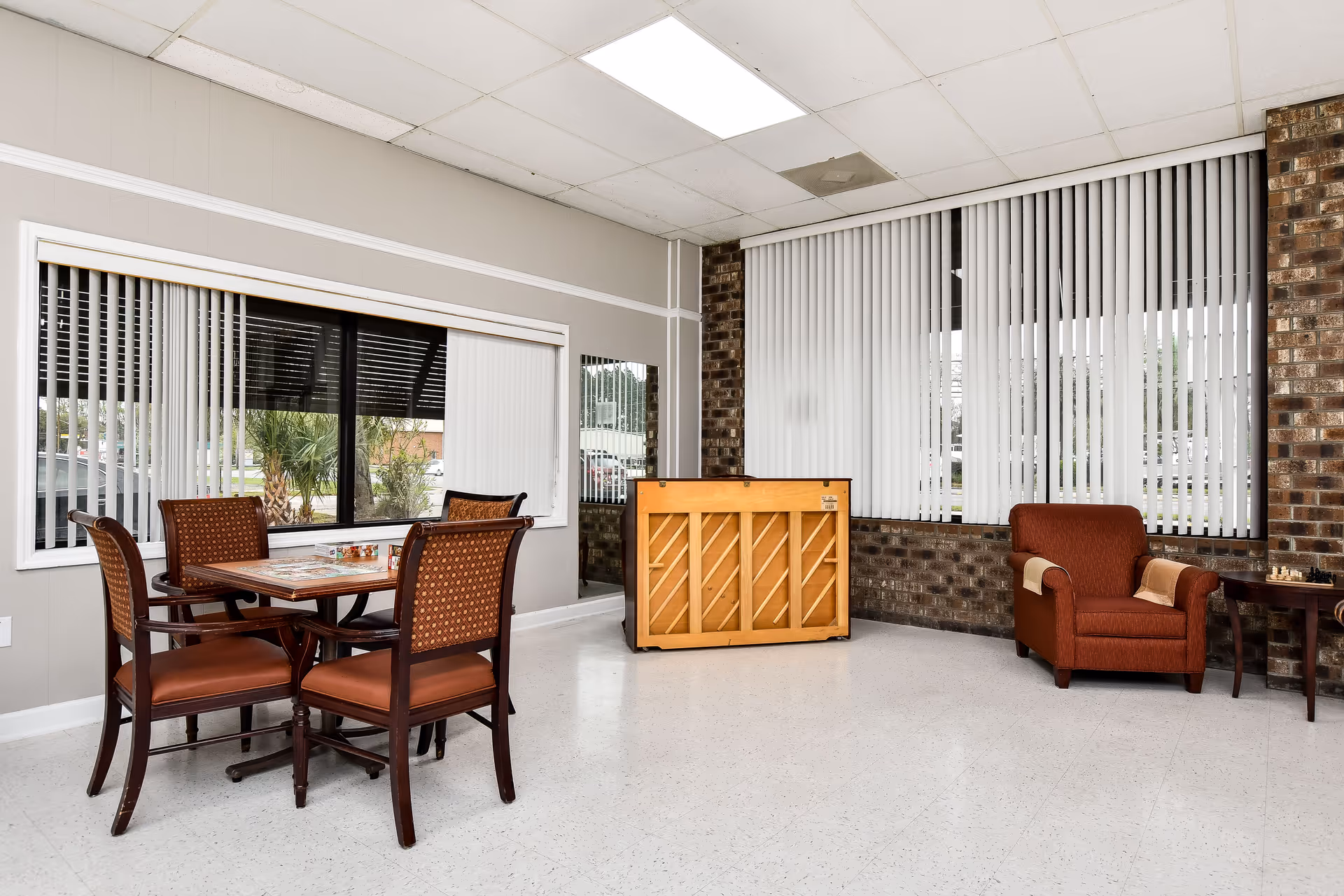 Bright indoor common area with a dining table, four chairs, piano, and red armchair.