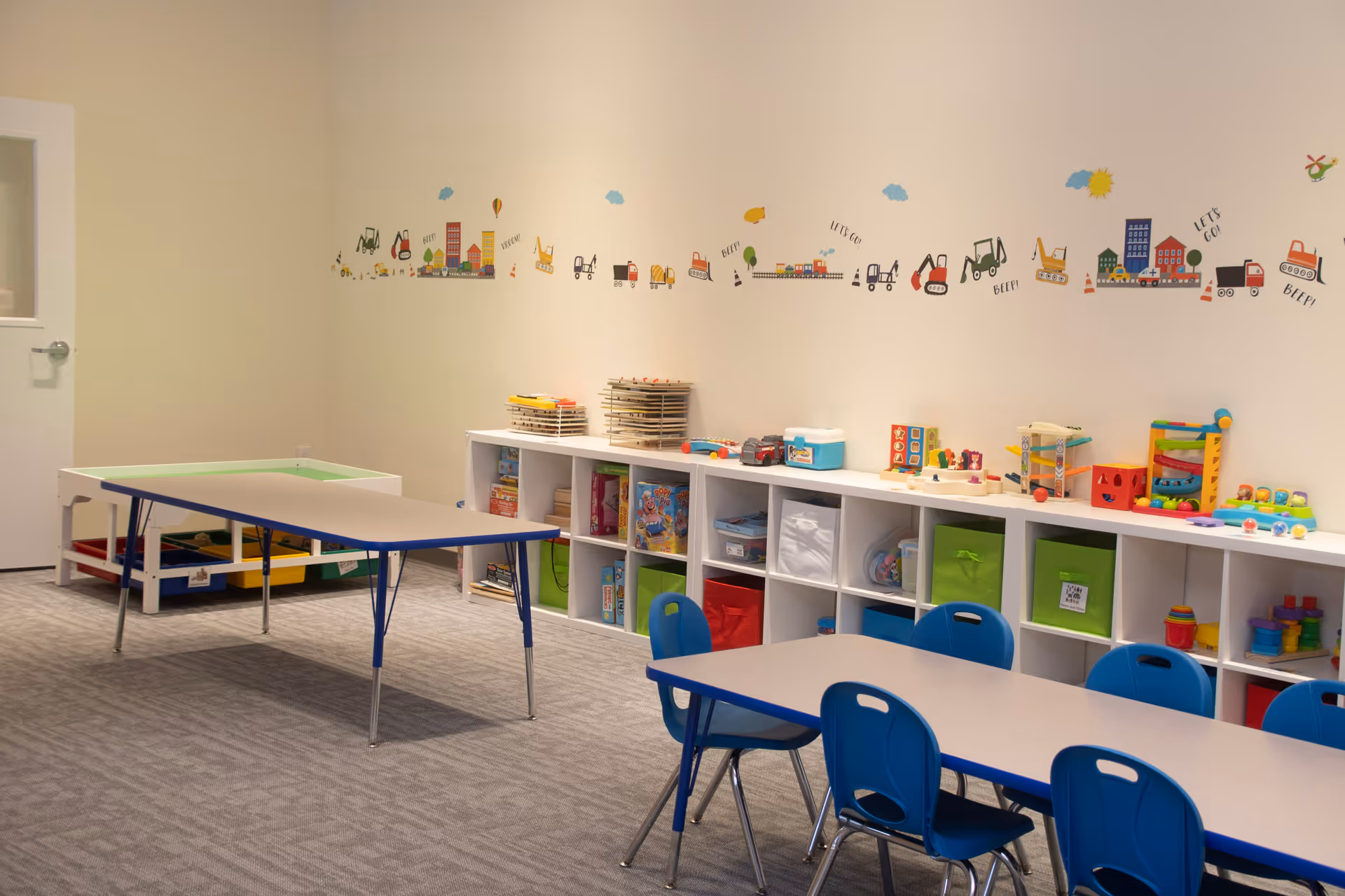 Bright pediatric activity room with tables, blue chairs, and toy storage along wall.