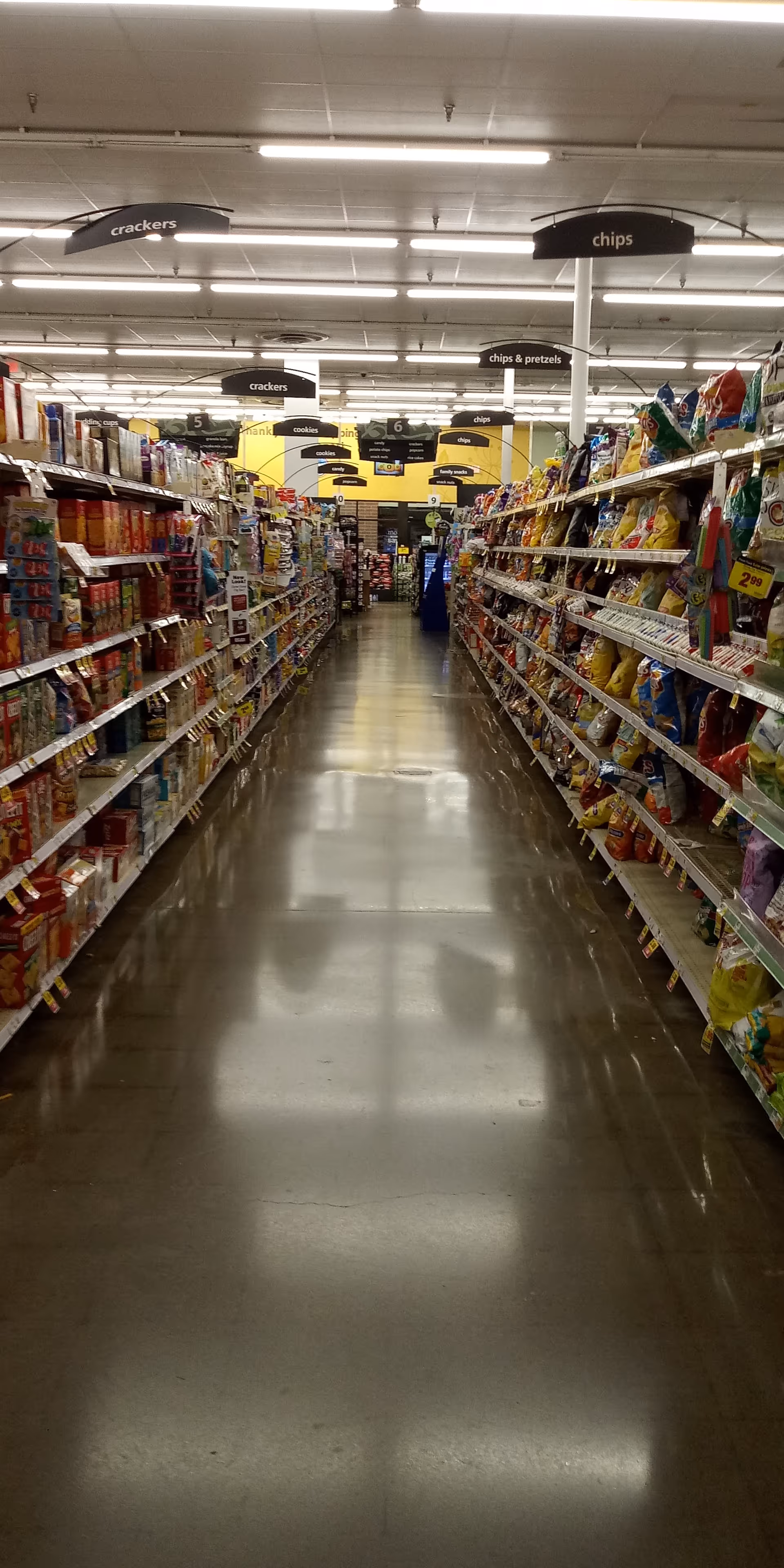 Long grocery store aisle with shelves stocked with snacks and overhead signs.