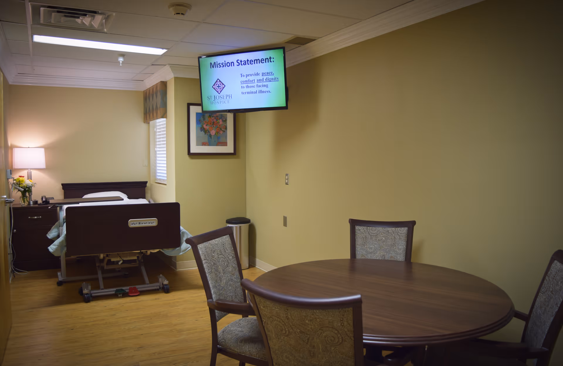A hospital bed beside a dresser and a round dining table in a hospice room.