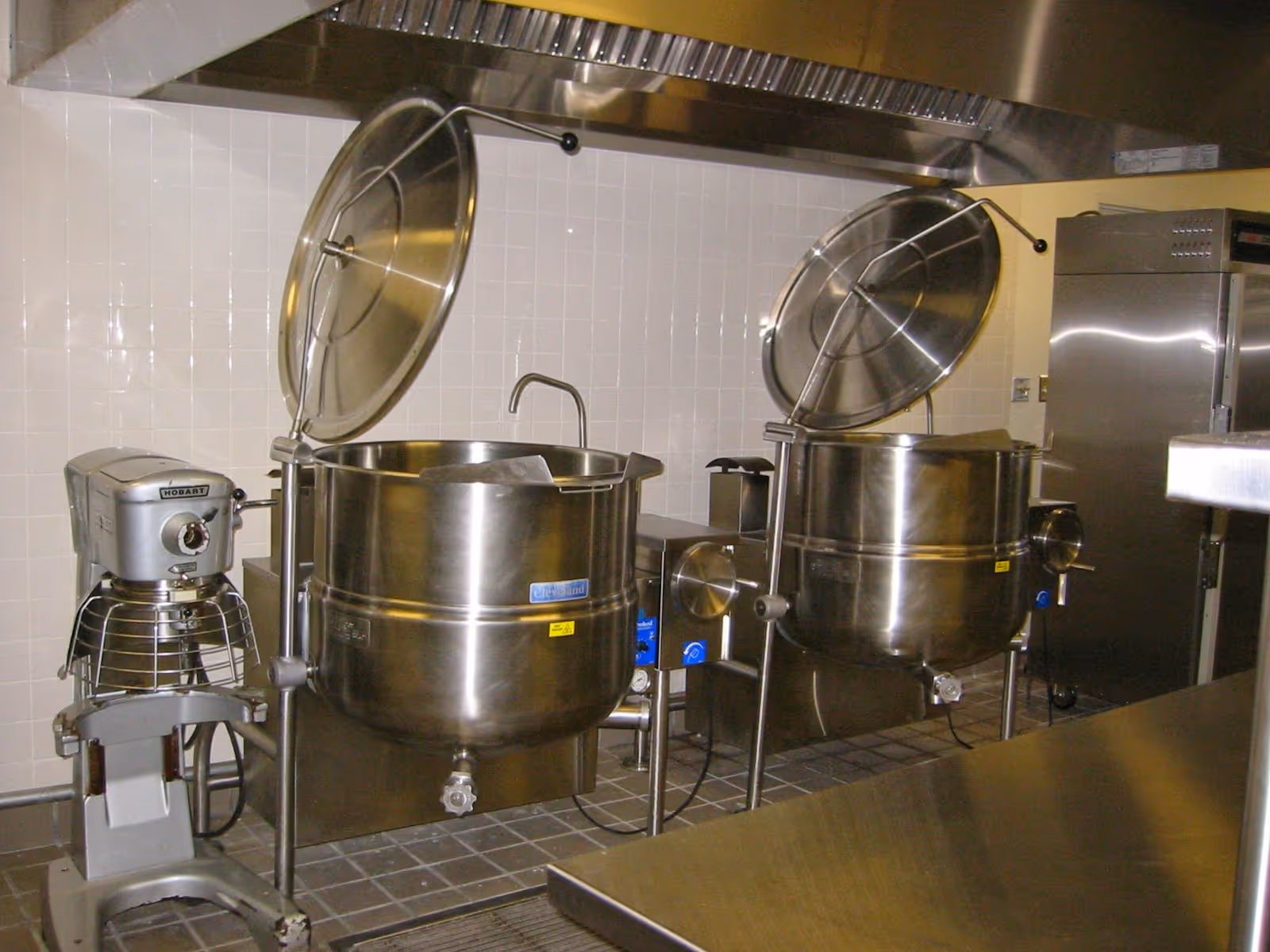 Industrial stainless steel kitchen with large kettles and mixers against tiled walls.