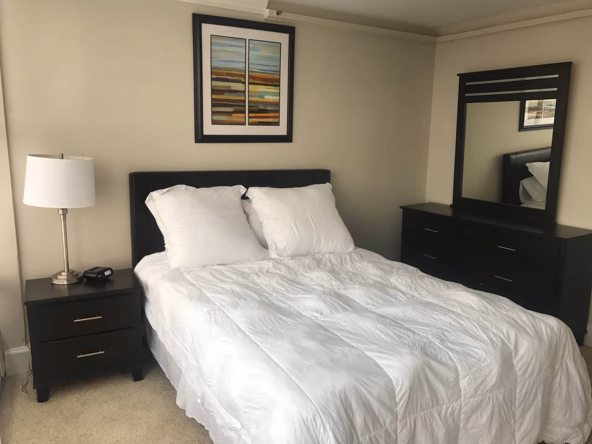 A neatly made bed with white bedding in a furnished bedroom, with dresser, lamp, and artwork.