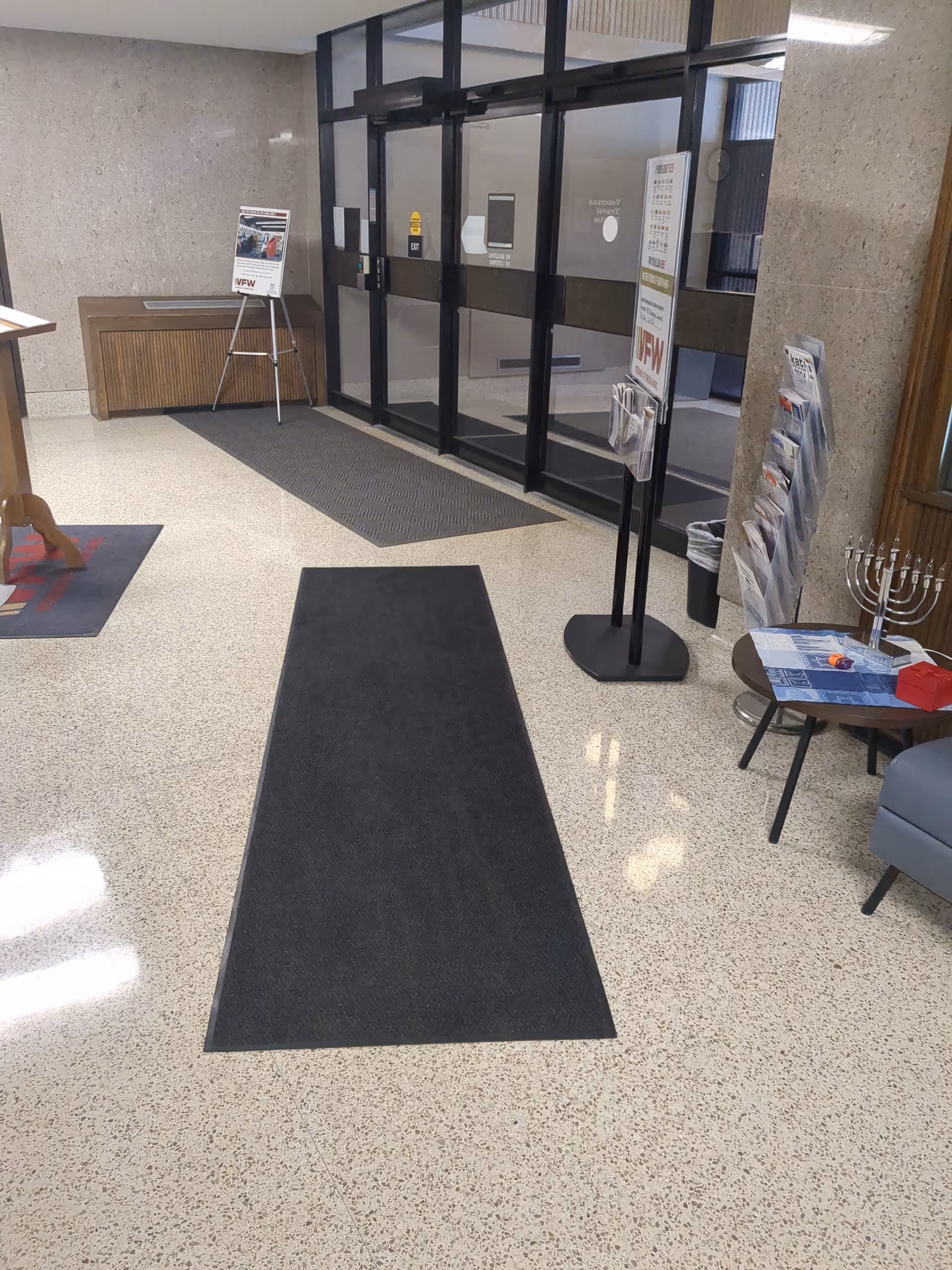 Interior lobby area with glass doors, mats, magazines, and a small reception table.