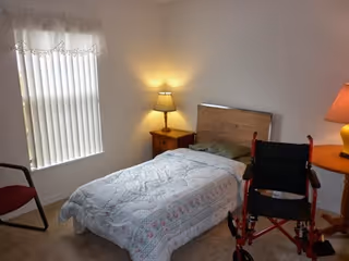 Bedroom with a single bed, two lamps, nightstands, window blinds, and a wheelchair beside the bed.