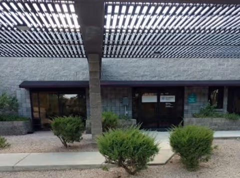 Exterior view of a facility entrance with a shaded overhead lattice and greenery.