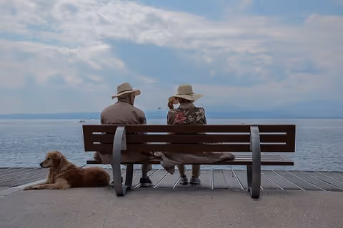 Two people sit on a bench by the water, with a dog nearby.