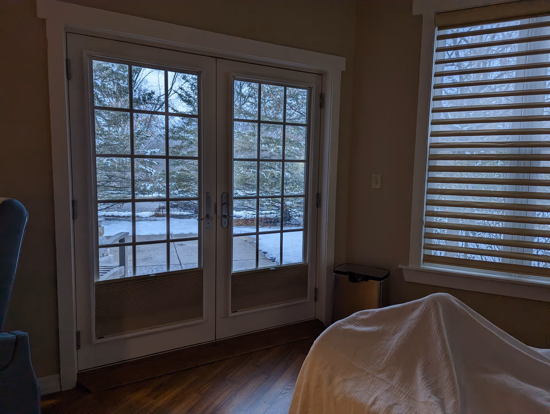 A dim bedroom with French doors opening to a snowy outdoor scene and a bed.