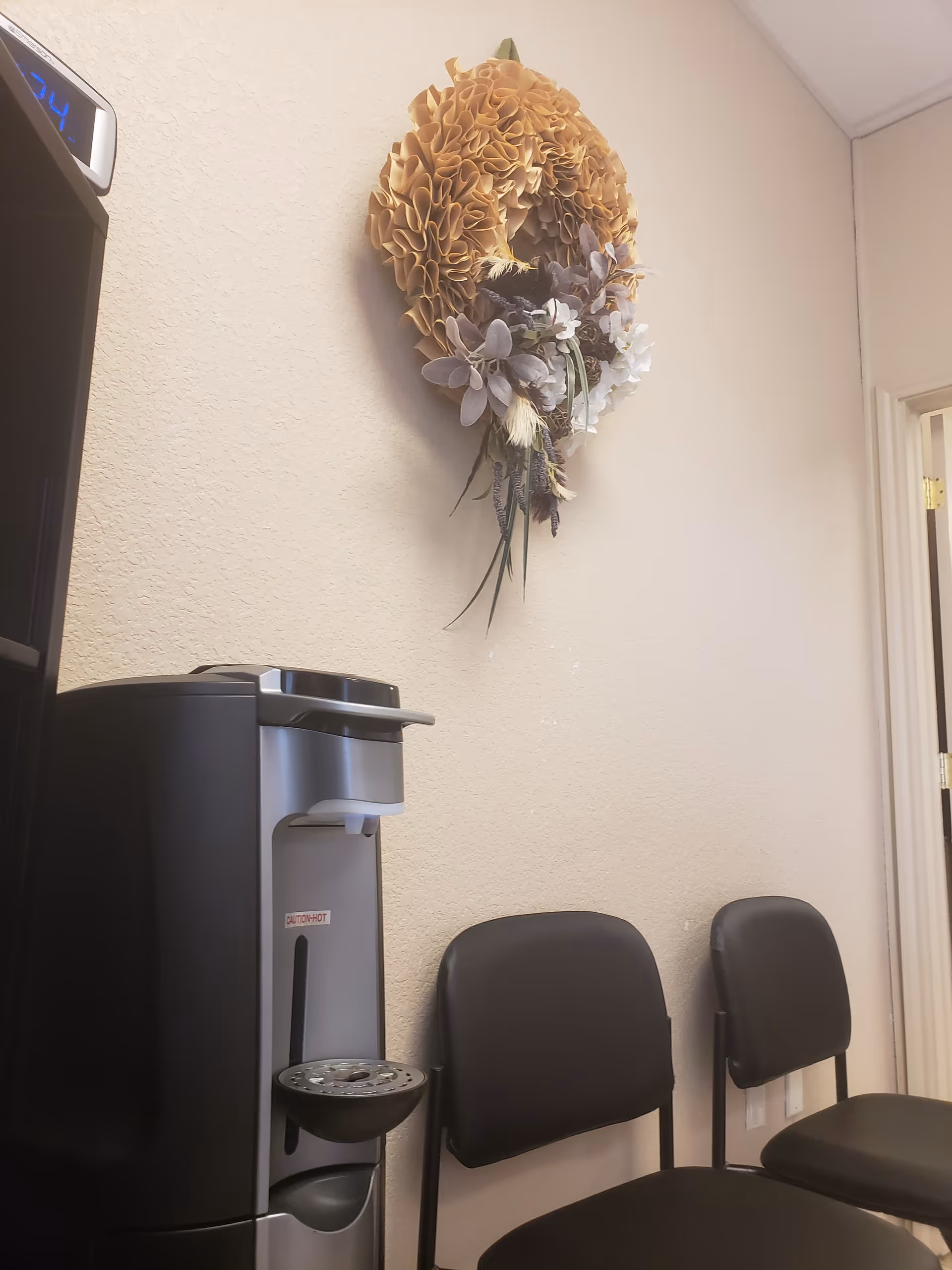 Interior corner with coffee dispenser, black chairs, and decorative wreath on beige wall.