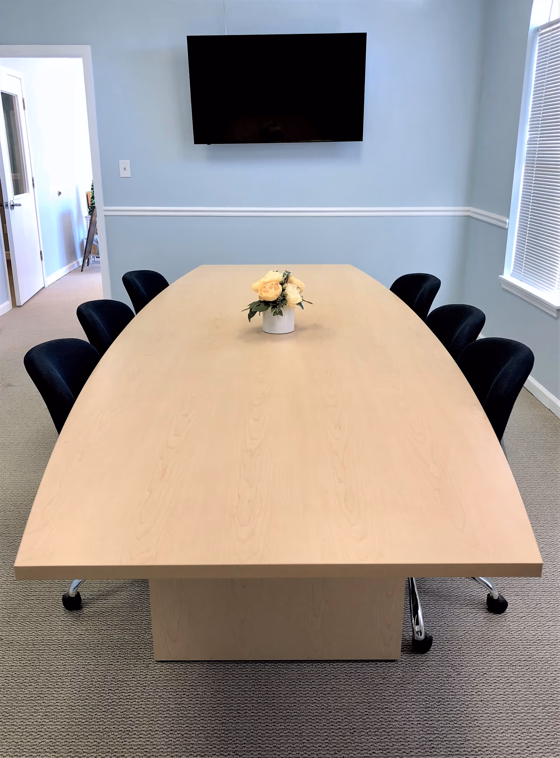 A light wood table with black chairs, floral centerpiece, and a wall-mounted TV.