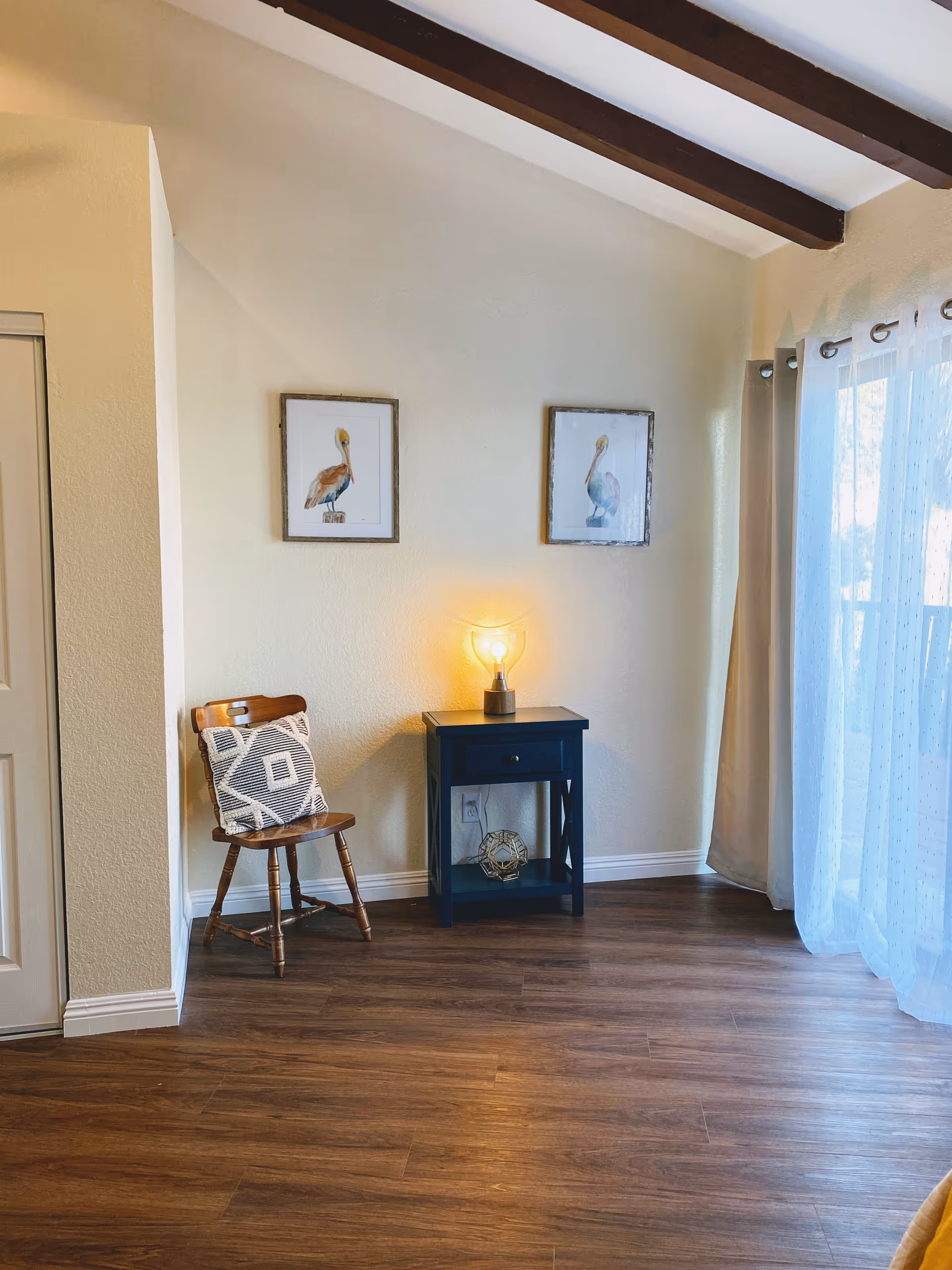 Cozy interior corner with a chair, small black side table, lamp, artwork, and curtains.