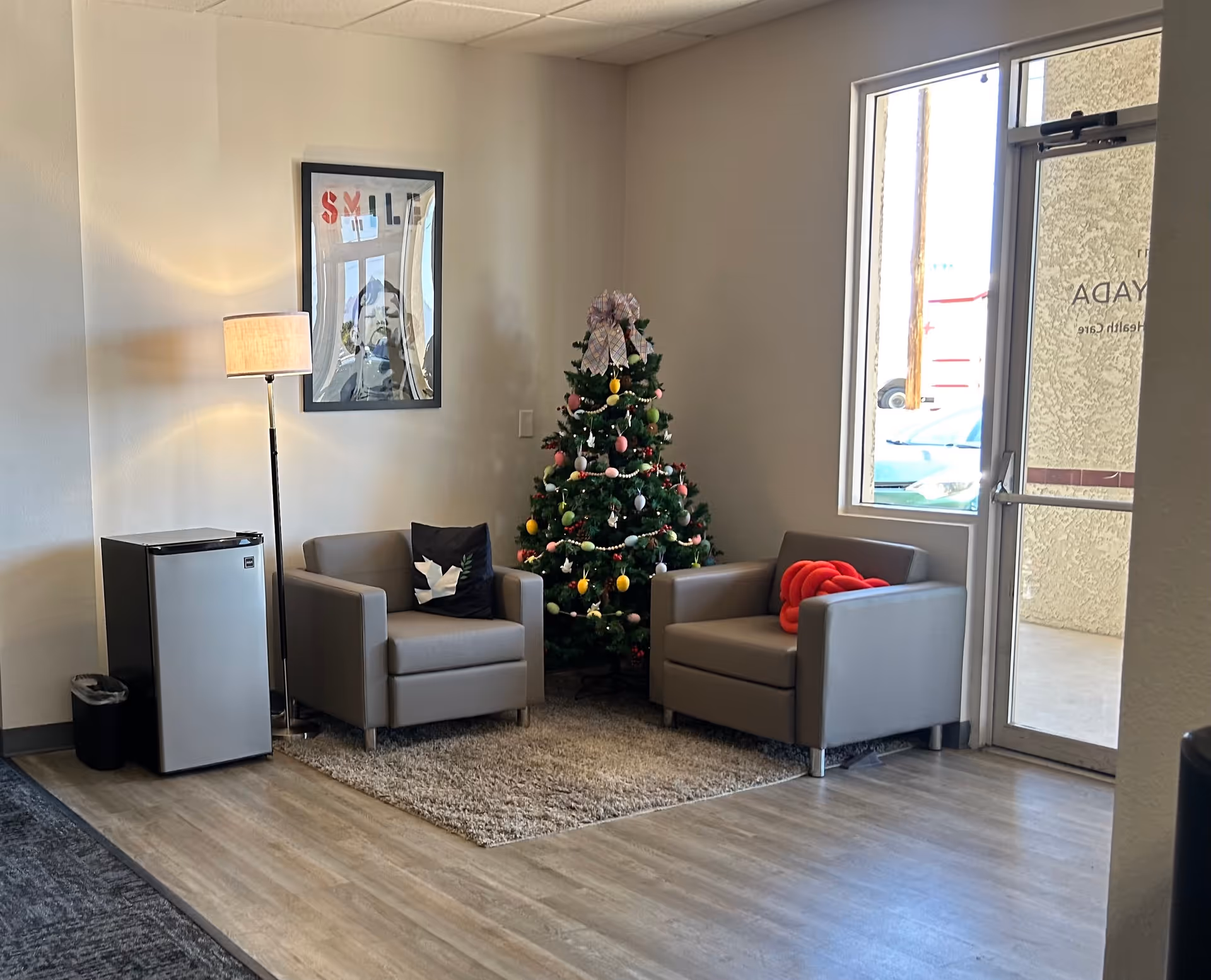 Cozy interior lounge area with a Christmas tree, two gray armchairs, a lamp, and a doorway.