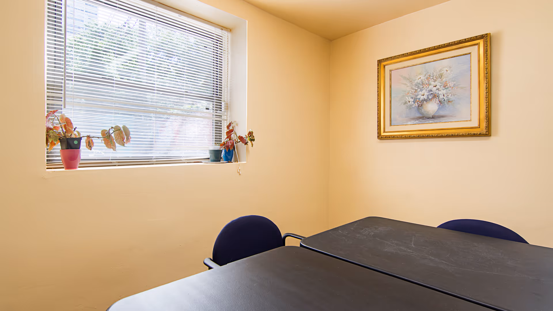 A small beige dining area with window, plants, framed artwork, a table, and blue chairs.