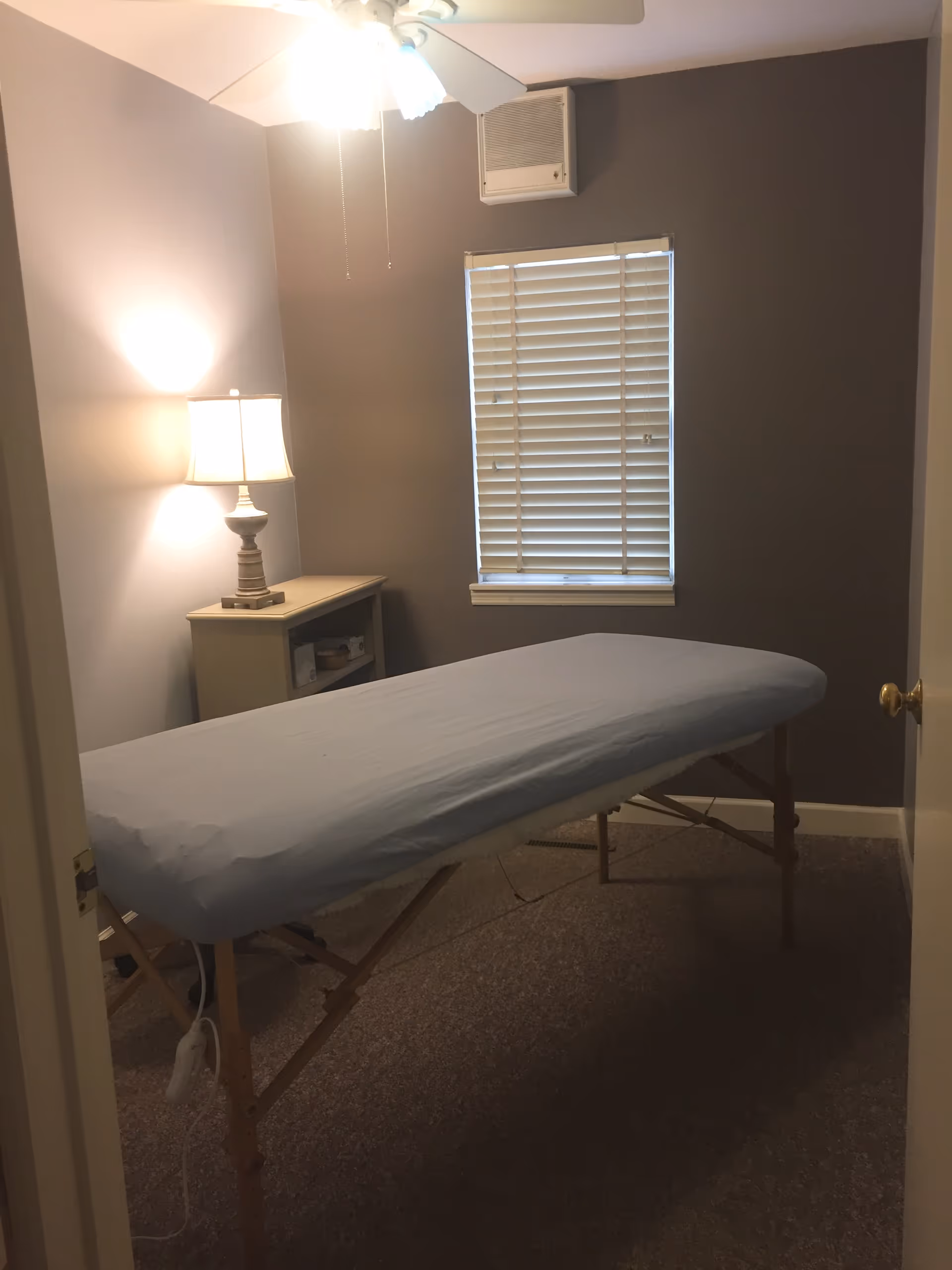 An empty chiropractic exam table in a small gray room with a window and lamp