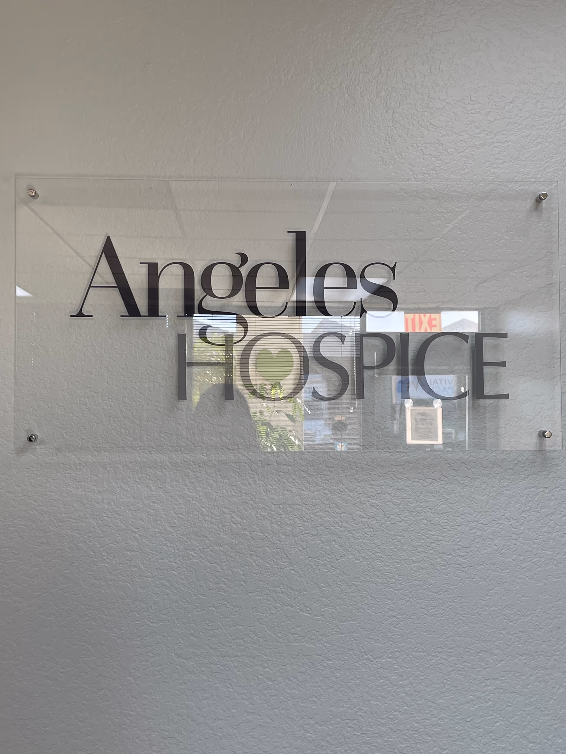 Clear glass sign reading Angeles Hospice mounted on a textured light gray interior wall.