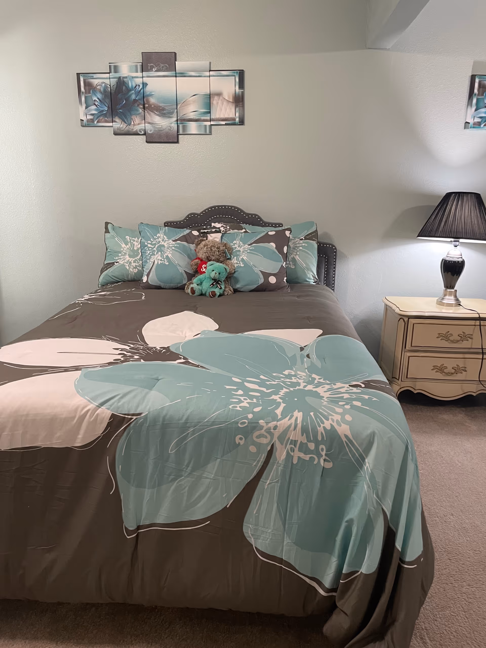 A neatly made bed with teal floral bedding, pillows, stuffed animals, and a bedside lamp.