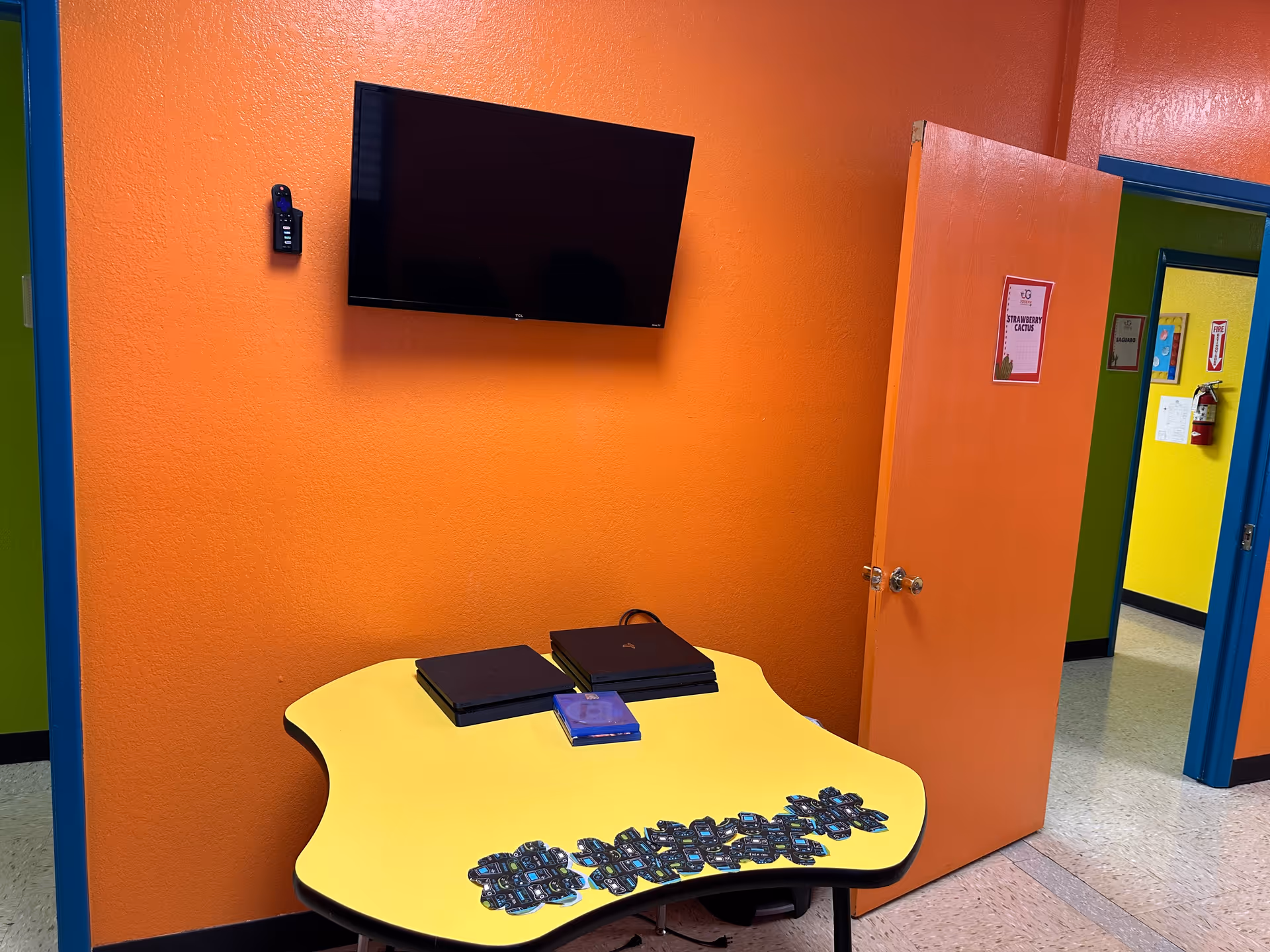 Bright orange wall, yellow curved table with numerous cards, and a wall-mounted flat-screen TV.