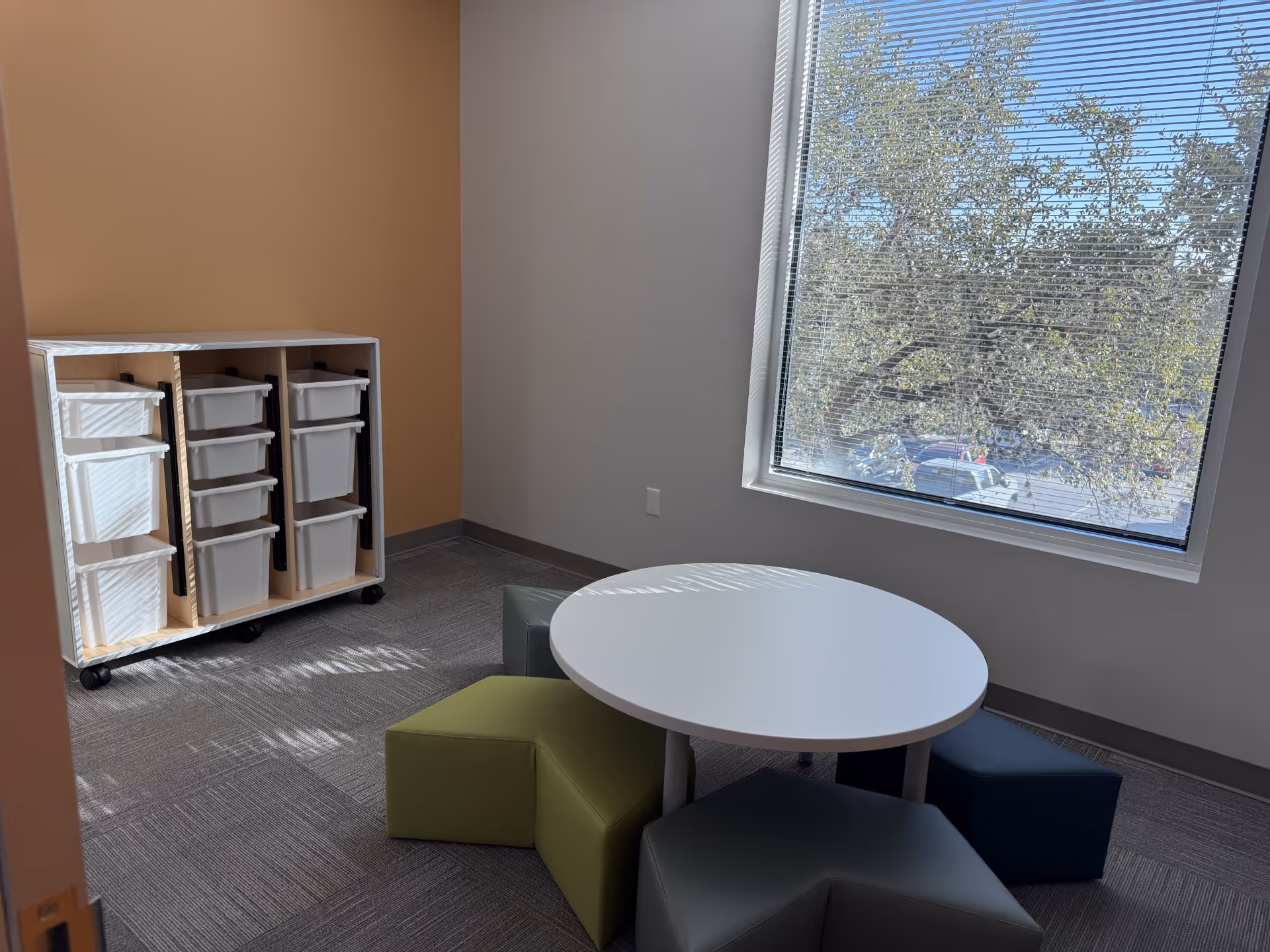 Indoor lounge area with round table, colorful ottomans, and storage carts beside a window.