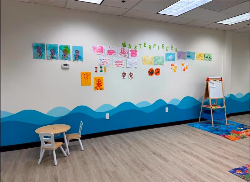 Bright therapy room with blue wave mural, children's artwork, a small table, and easel.