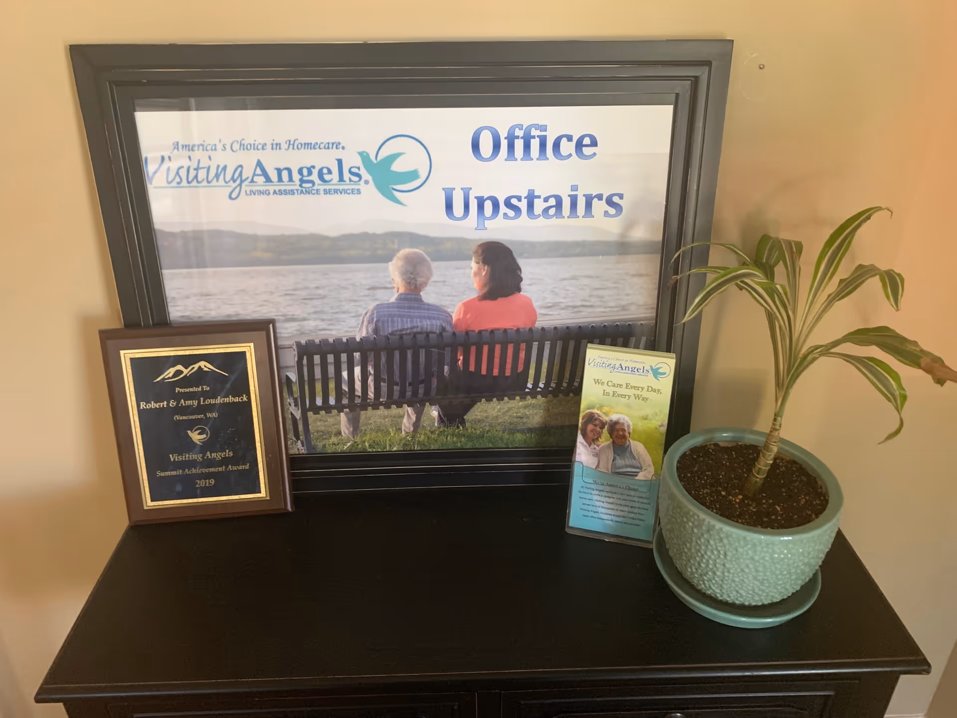 Framed Visiting Angels sign with bench scene, a plaque, brochure, and potted plant on a table