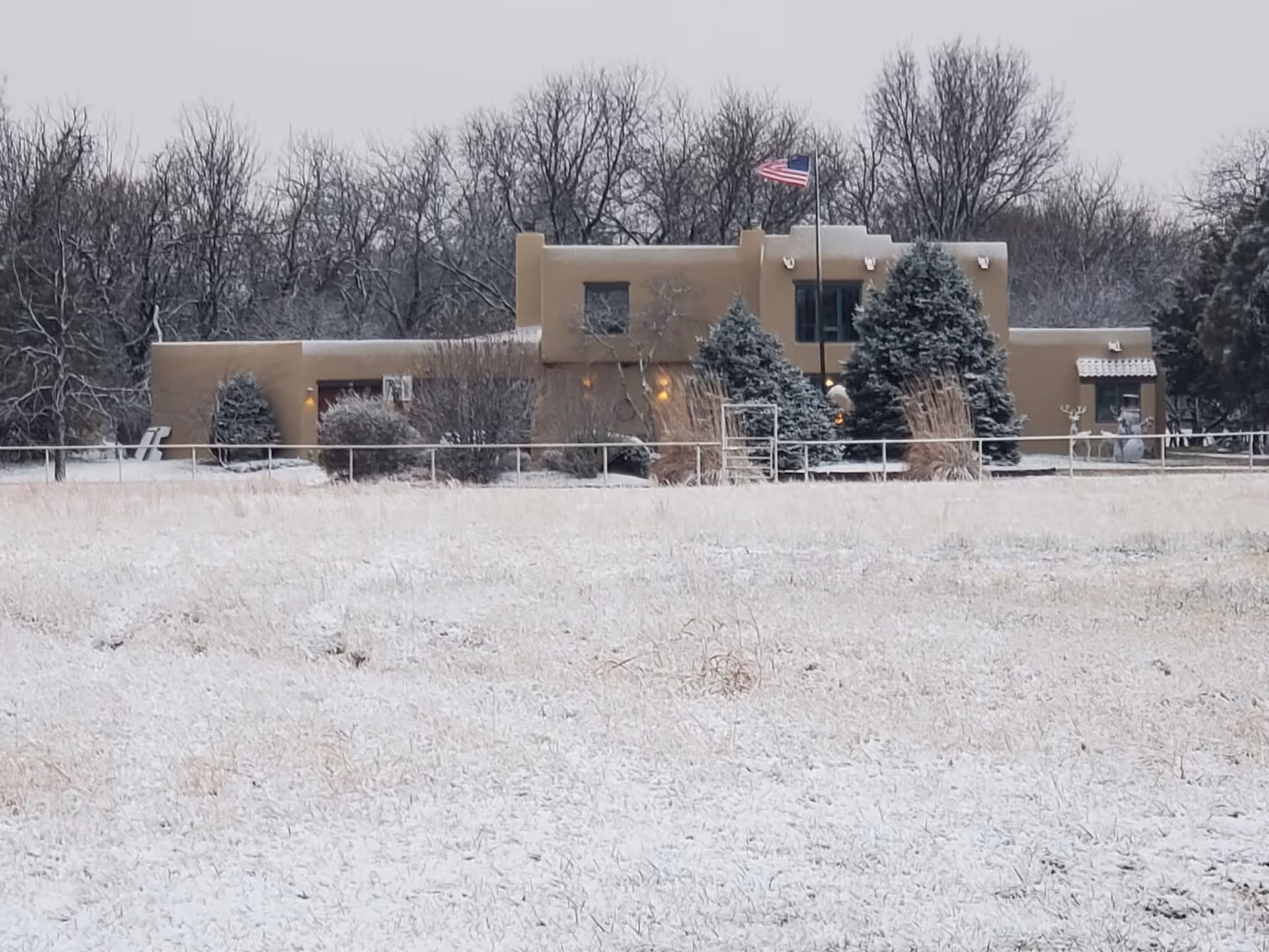 A tan flat-roofed building with an American flag, trees, and snow in front.