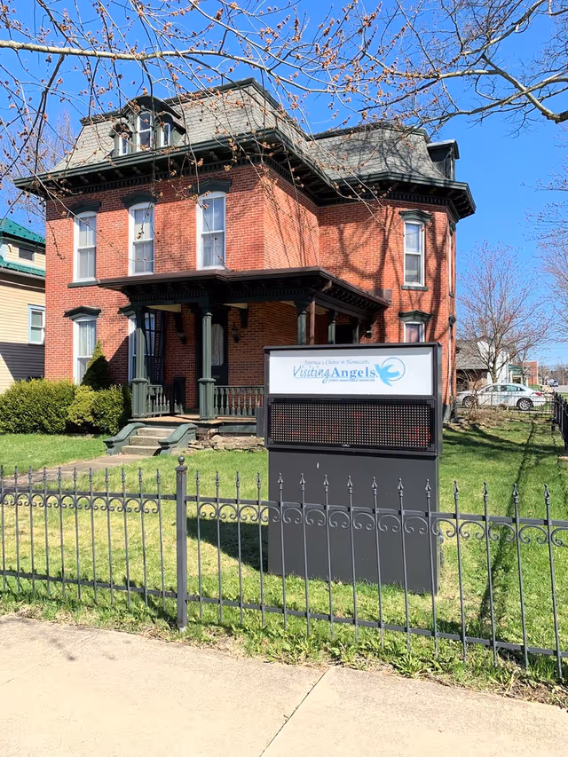 Brick two-story Visiting Angels care facility with sign, iron fence, and green lawn.