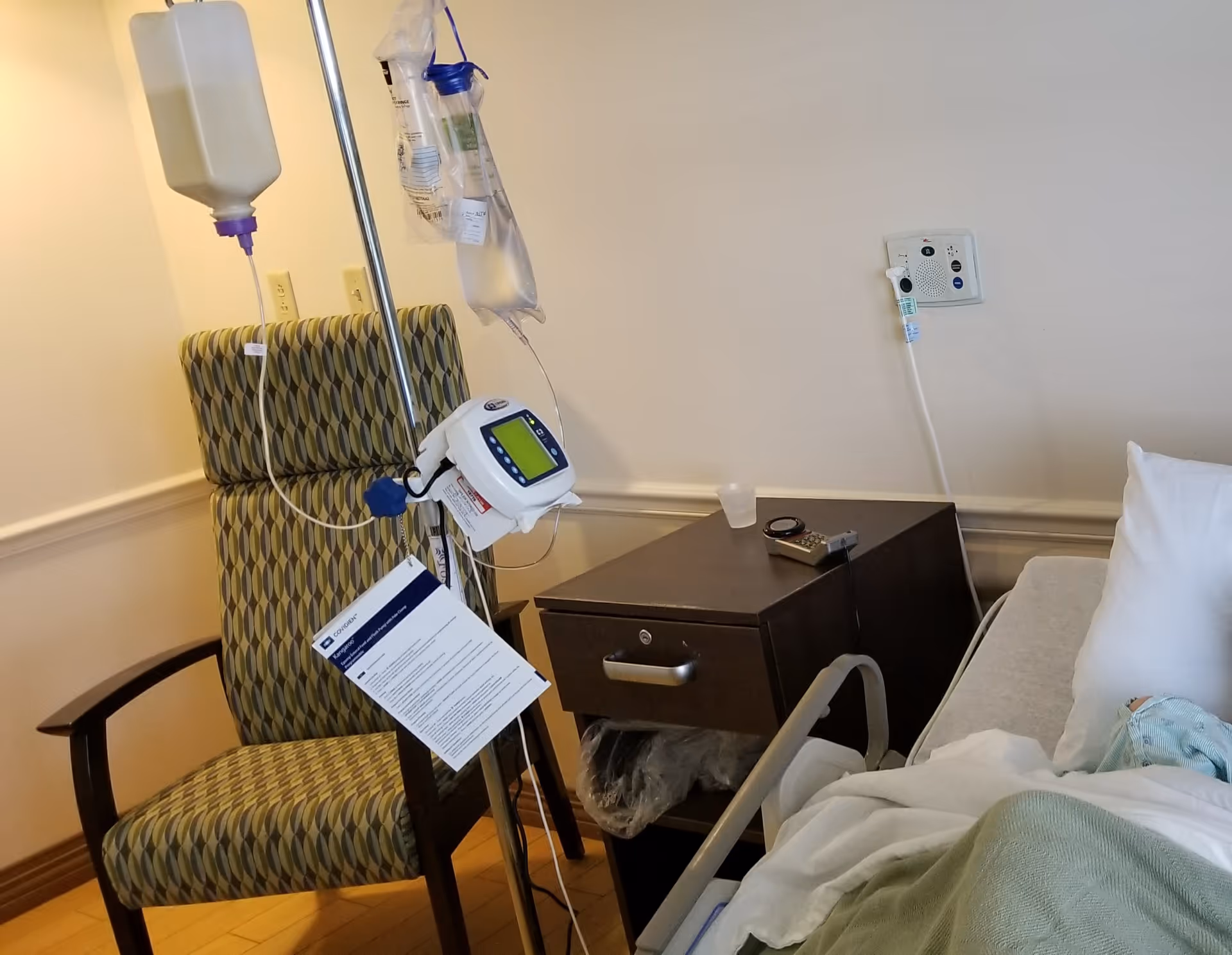 Hospital-style patient room with IV pole, infusion pump, and bedside table beside a bed.