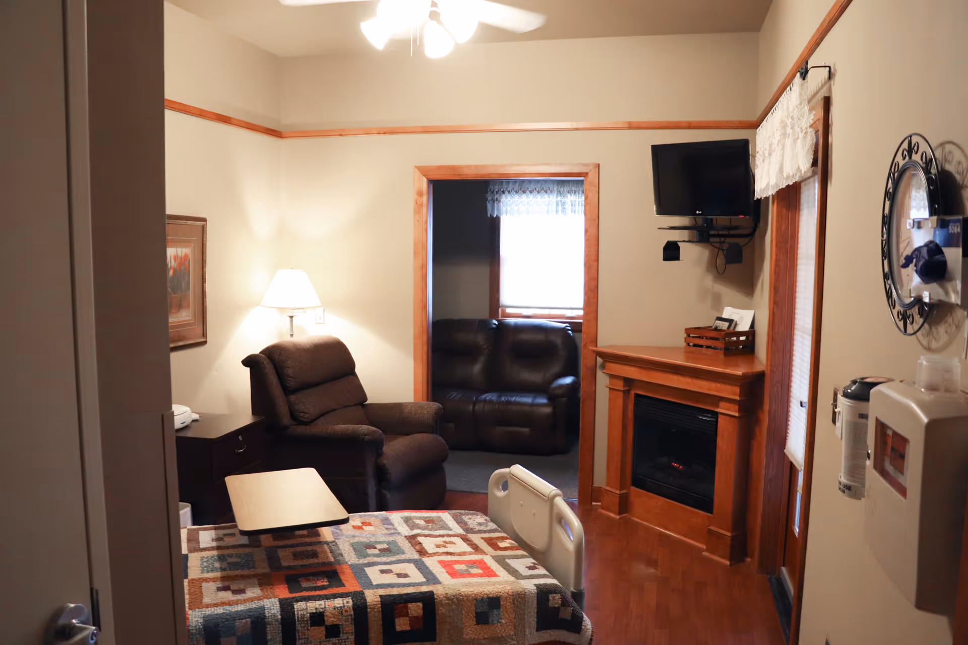 Cozy hospice living area with a quilted bed, recliner, fireplace, and wall-mounted TV.