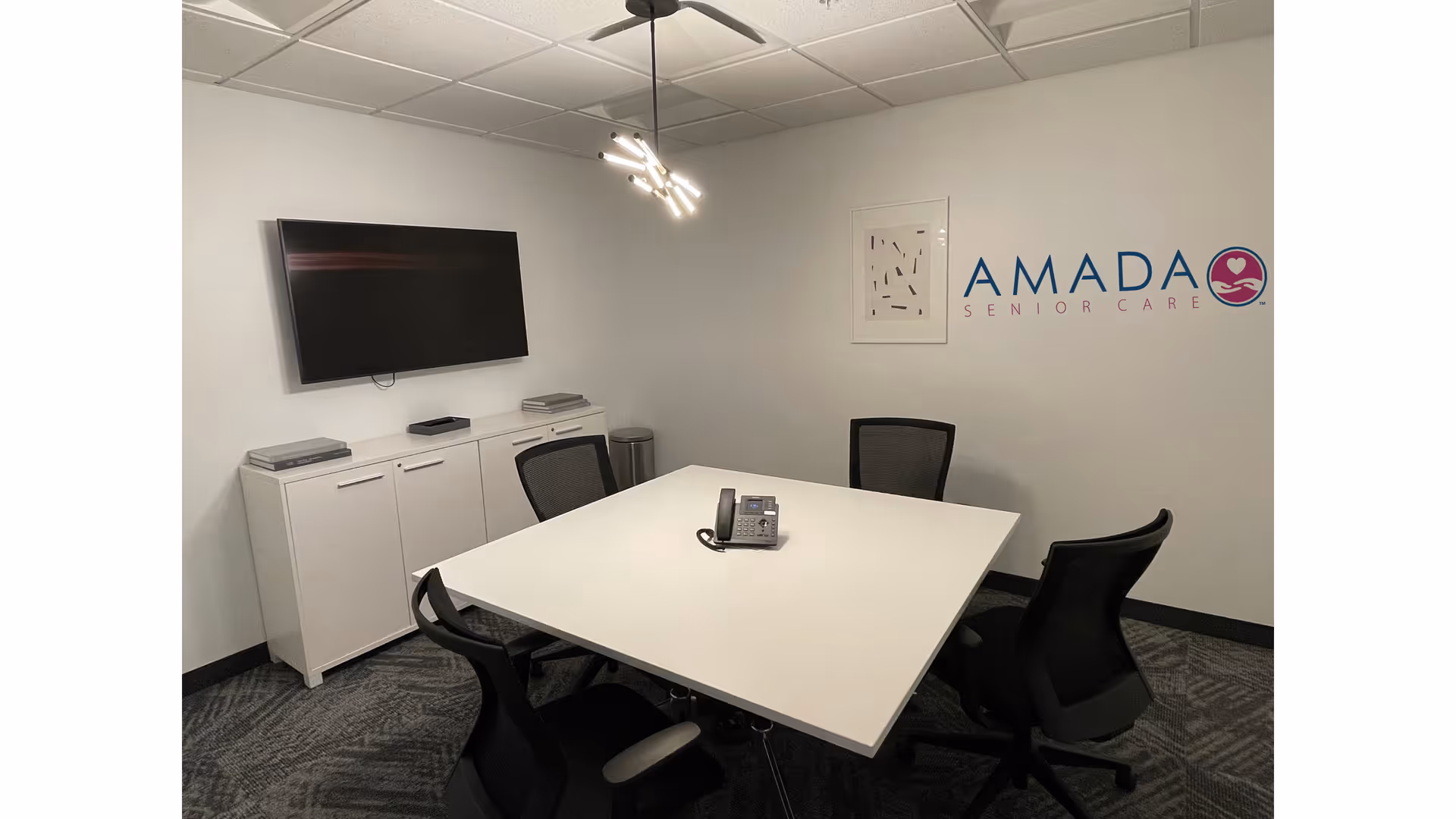 Modern conference room with white table, black chairs, wall TV, and Amada Senior Care logo.