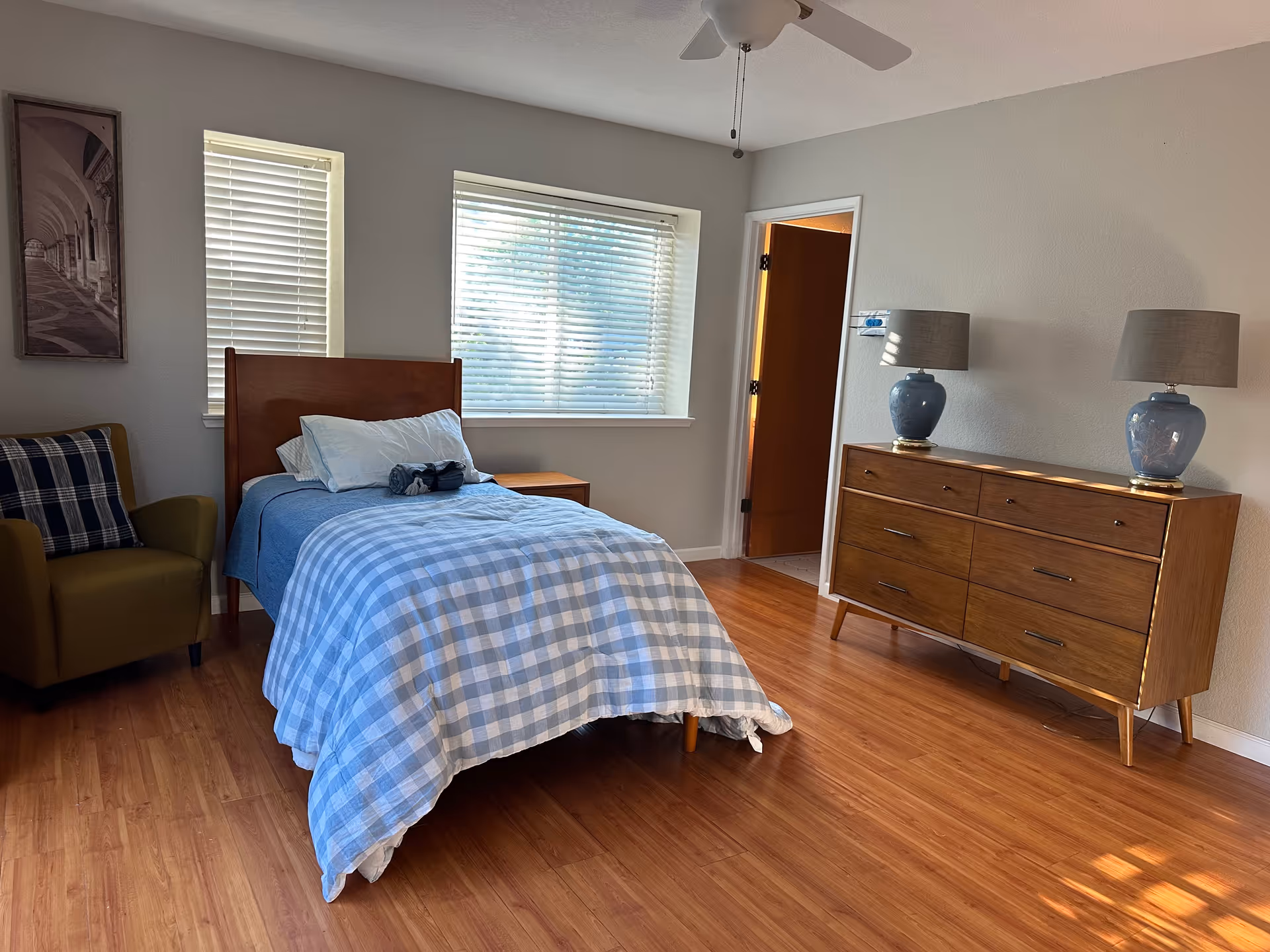Bedroom in a senior living facility with a bed, dresser, armchair, and two lamps.