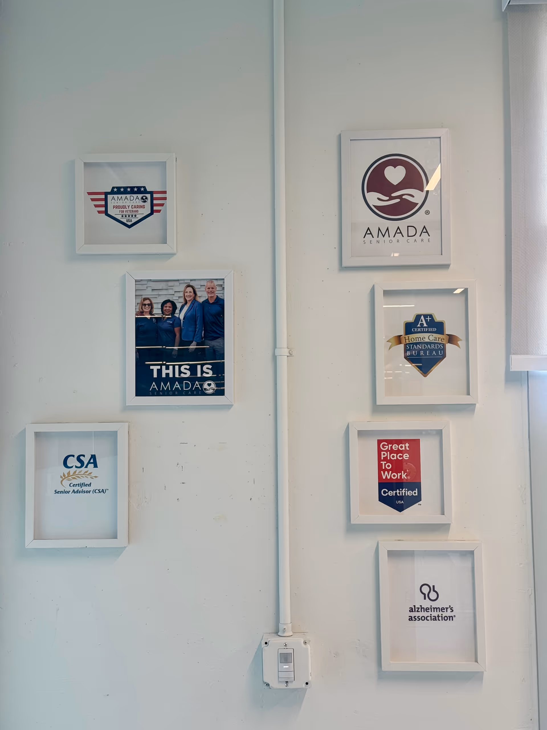 Interior wall displaying multiple framed Amada Senior Care logos and accreditation posters.