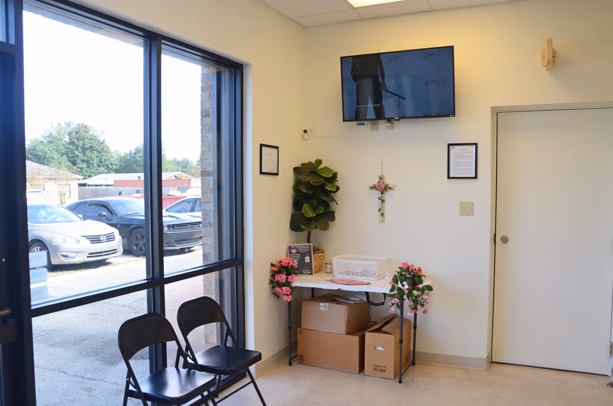 Bright entry area with large window, desk, potted plants, chairs, wall cross, and wall-mounted TV.