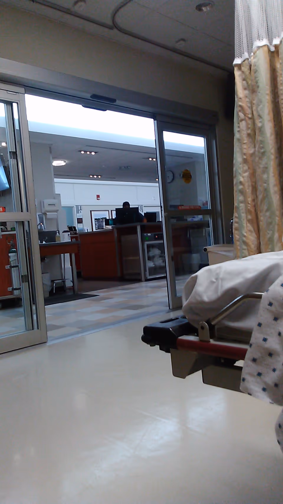 Interior hospital corridor with sliding doors and a patient bed visible.