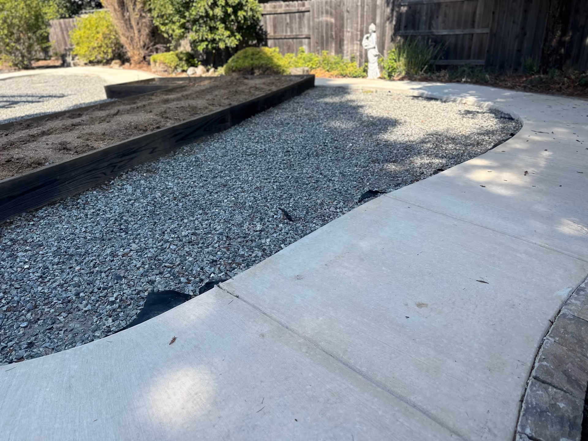 Curved concrete path beside a gravel bed and statue in a landscaped outdoor area.