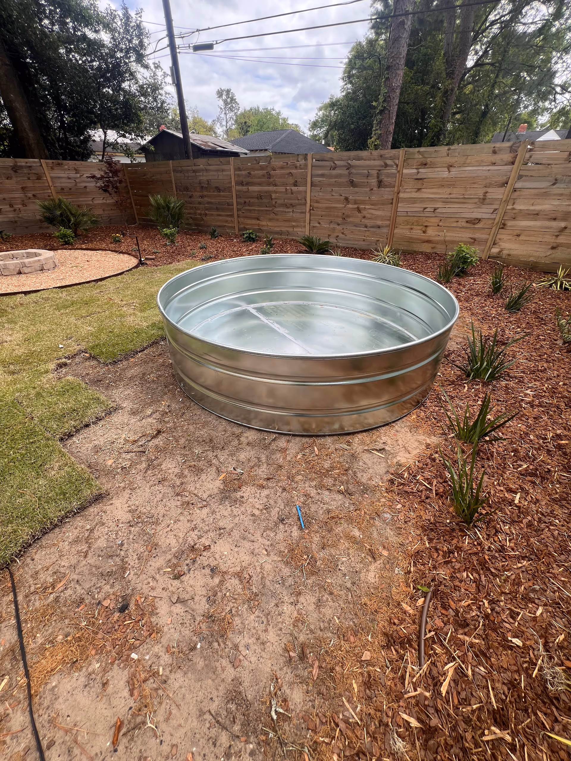 Round galvanized tub sits in a fenced backyard with mulch and plants.