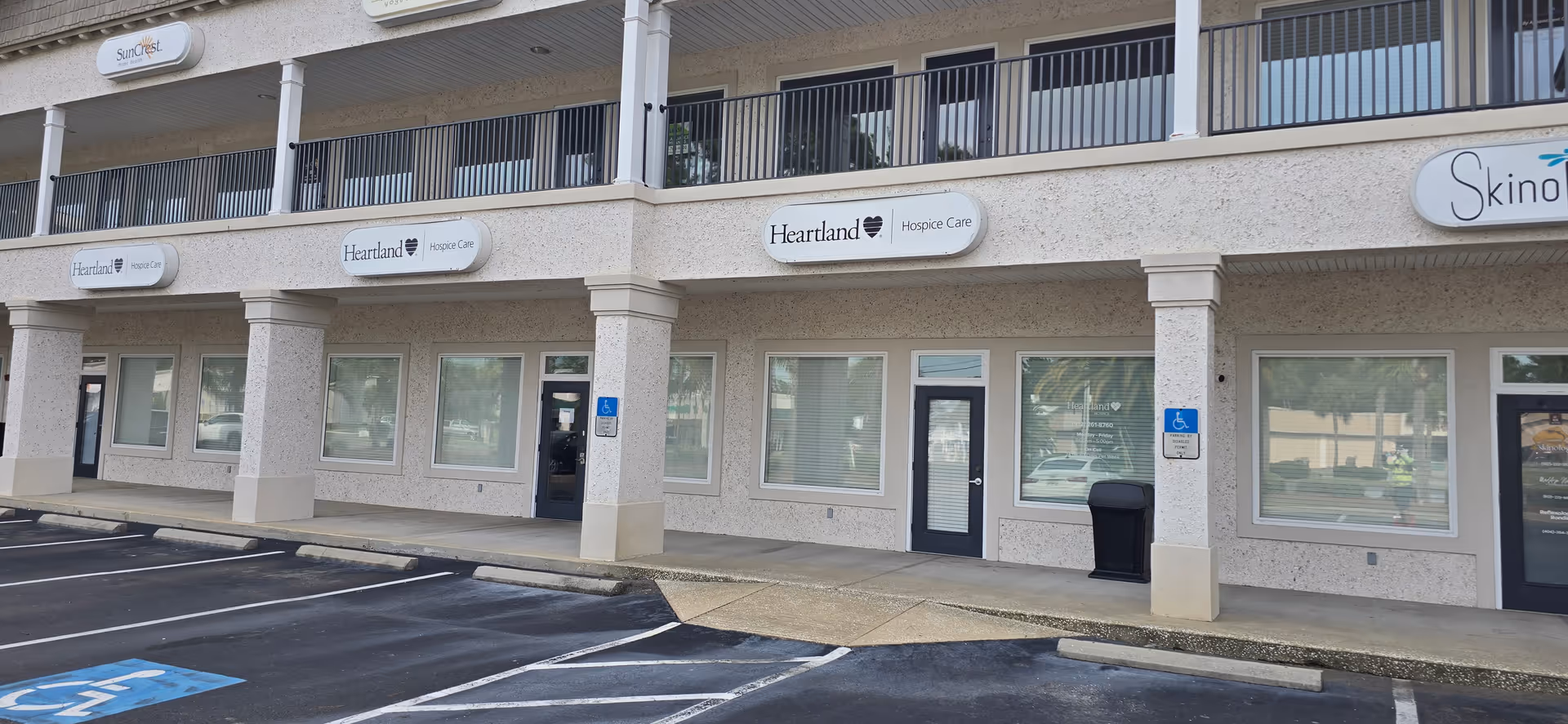 Facade of a Heartland Hospice facility with storefronts and a parking lot in front.