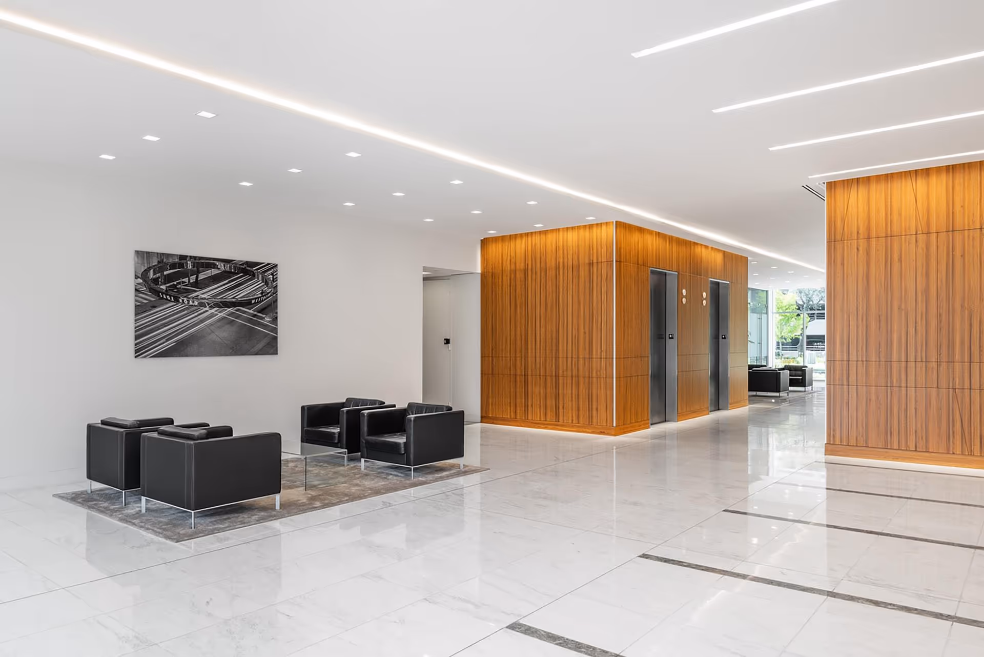 A bright modern lobby with white walls, black lounge chairs, marble floor, and wood-paneled elevators.