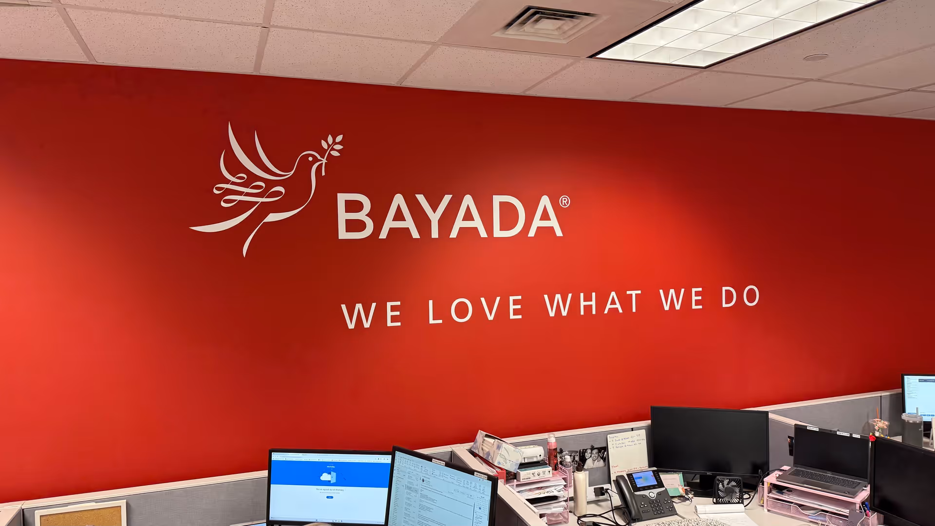 Red BAYADA wall with logo and slogan; office desks and computers in foreground.