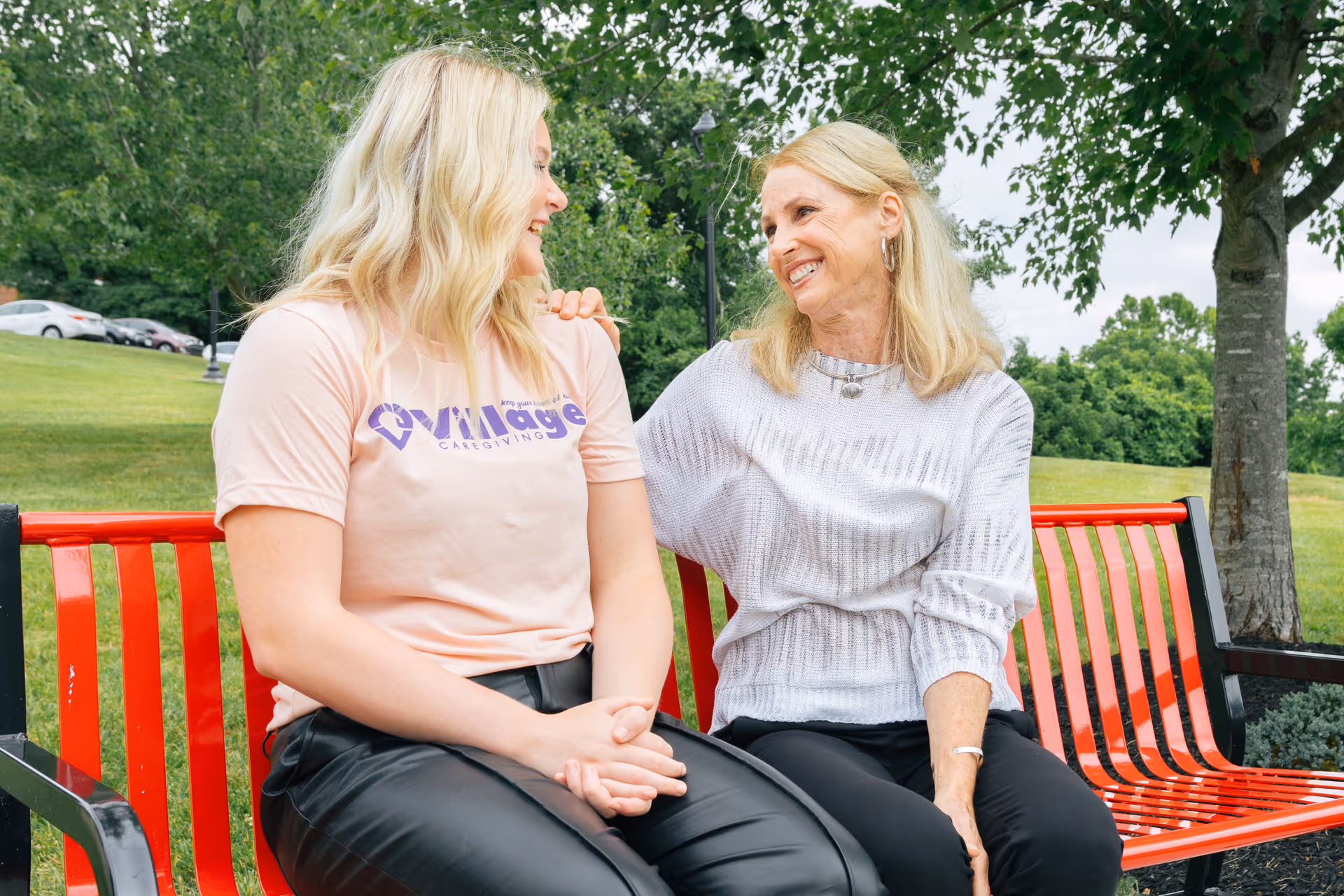 Two women sit on a red bench outdoors, smiling and talking.