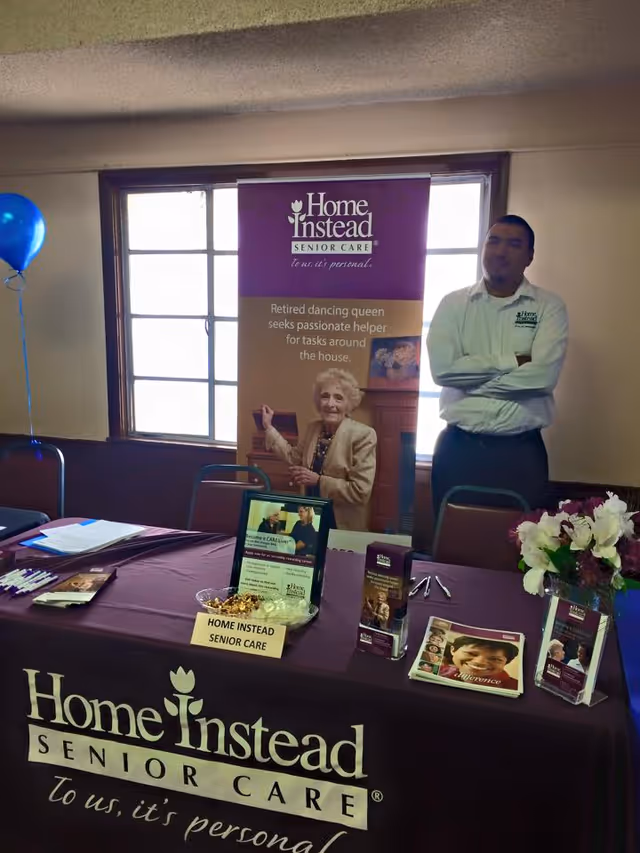Promotional table and banner for Home Instead Senior Care inside a facility, with staff nearby.