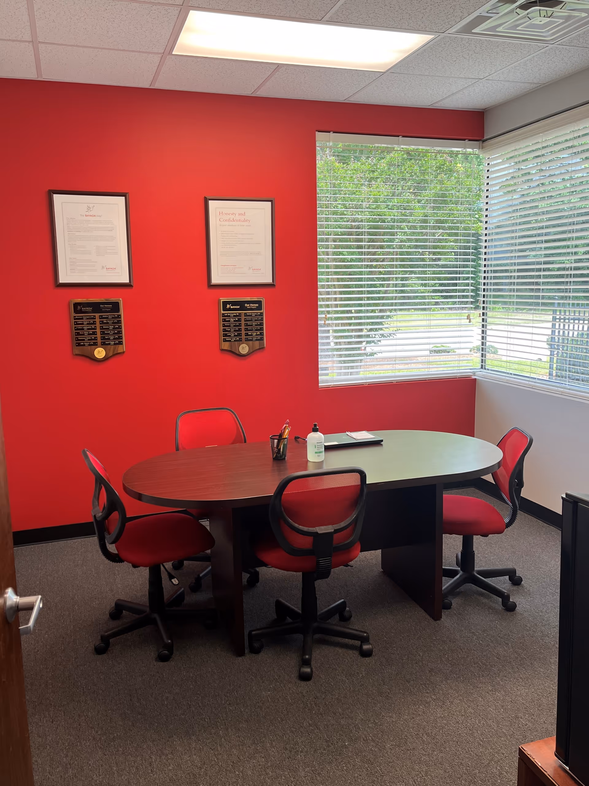 A red-walled meeting room with a round table, red chairs, and large window blinds.